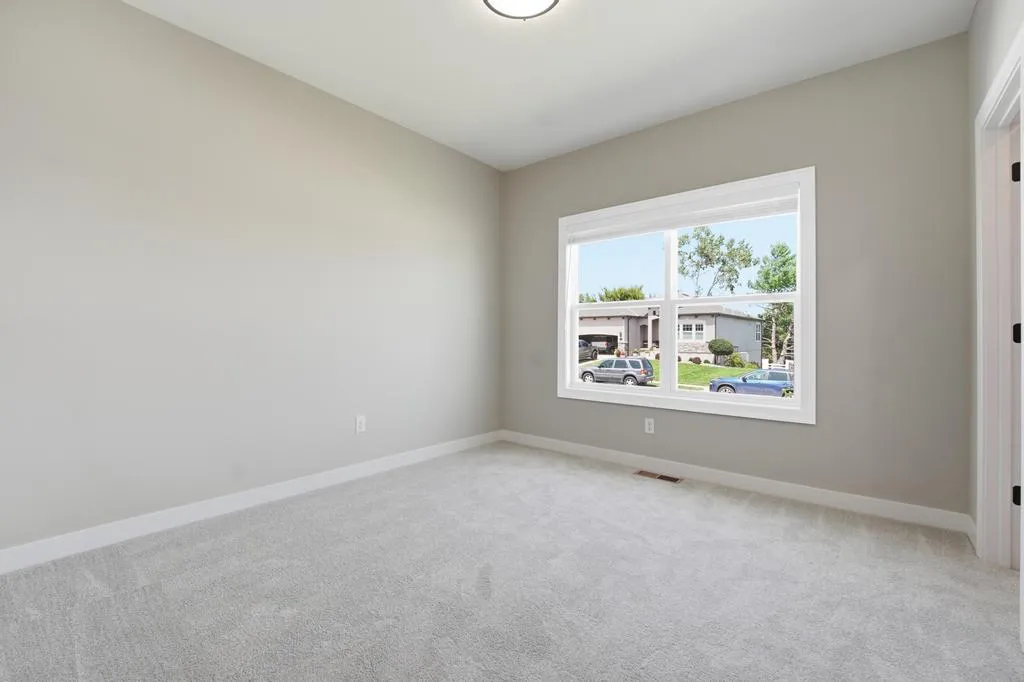 Empty bedroom with beige walls, carpeted floor, and window overlooking residential street with parked vehicles.