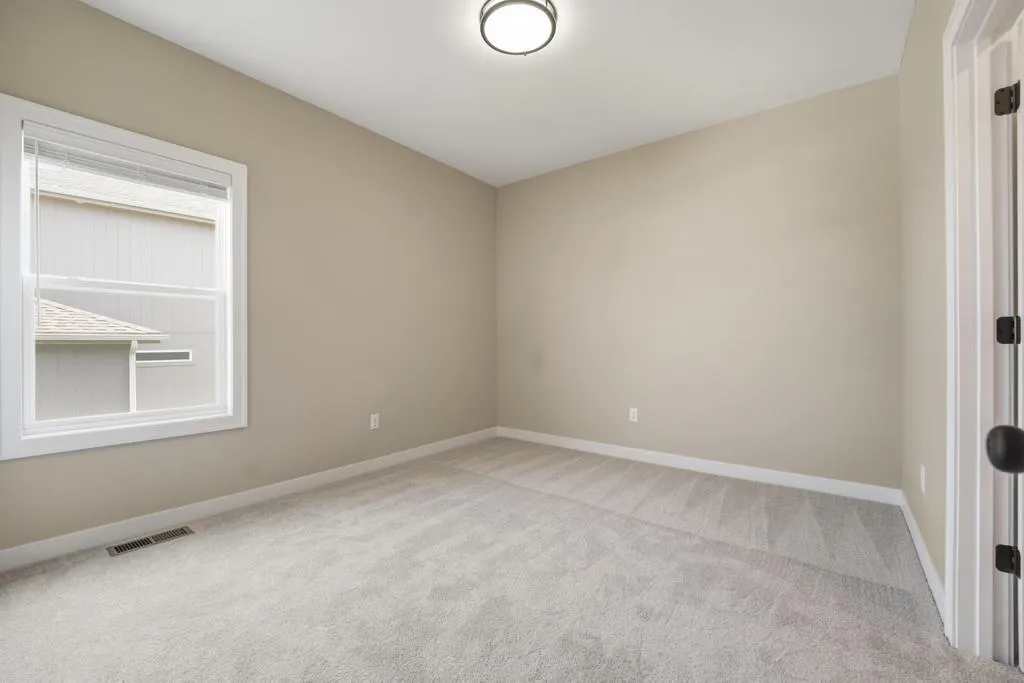 Empty bedroom with beige walls, gray carpet, window, ceiling light fixture, and white trim doorway.