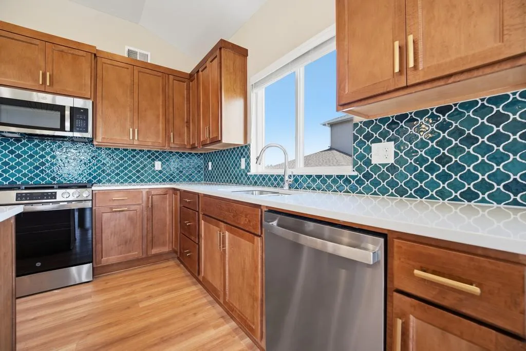 Kitchen with wood cabinets, teal patterned backsplash, white countertops, stainless steel appliances, and large windows.