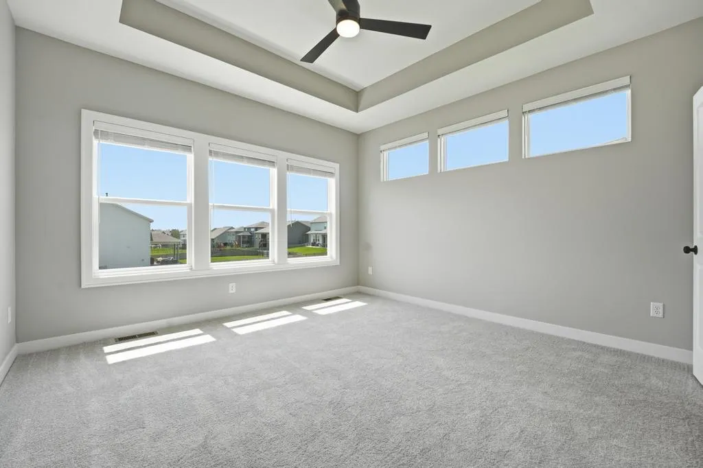 Empty room with gray carpet, gray walls, multiple windows with views of residential neighborhood, and black ceiling fan.