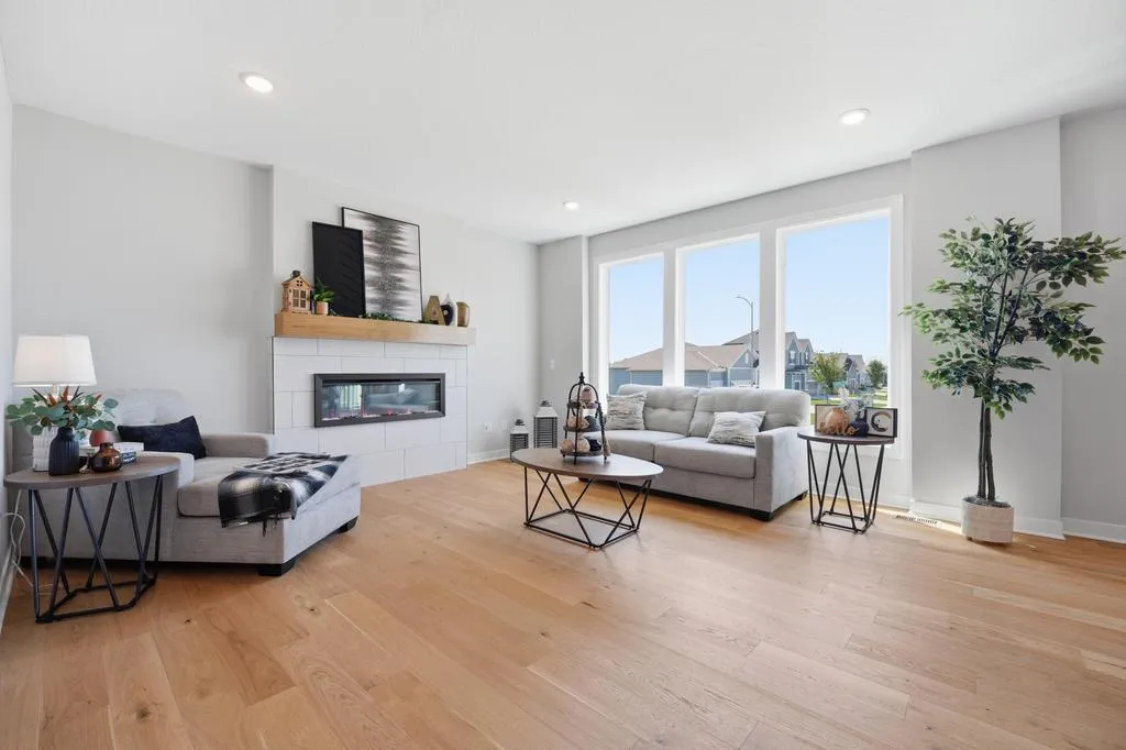 Living room with wood flooring, gray sofa, fireplace with white surround, and large windows overlooking residential