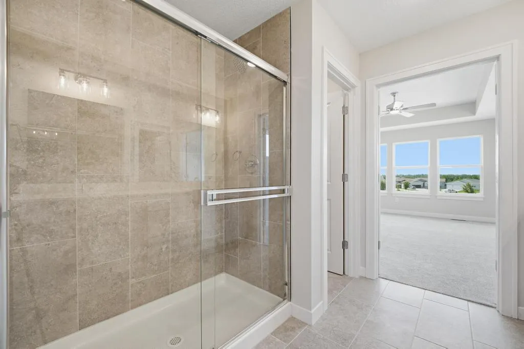 Bathroom with frameless glass shower enclosure, beige tile walls, and view of bedroom with windows overlooking landscape.