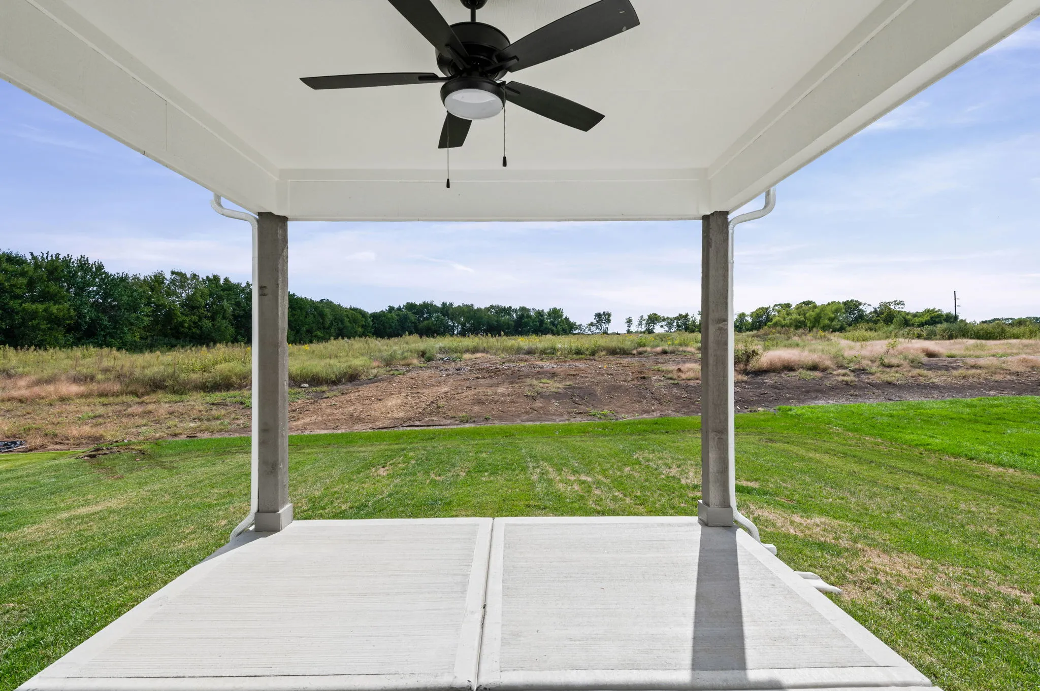 Covered patio with black ceiling fan overlooking green lawn and open field with tree line.