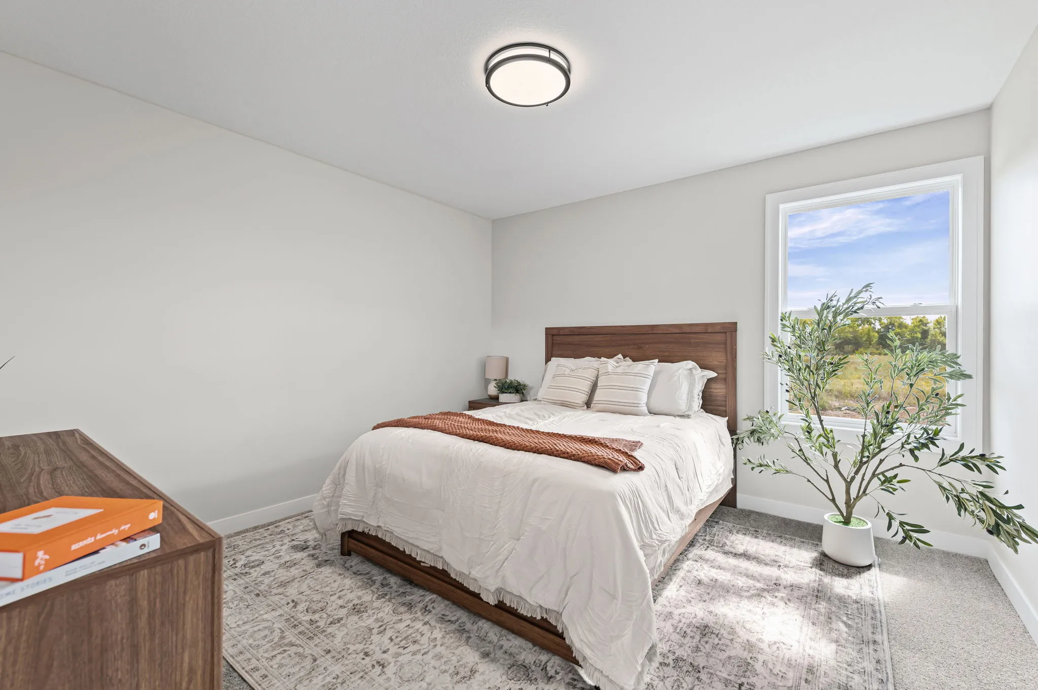 Bedroom with wood bed frame, white bedding, brown throw blanket, nightstand with lamp, dresser with orange boxes, potted