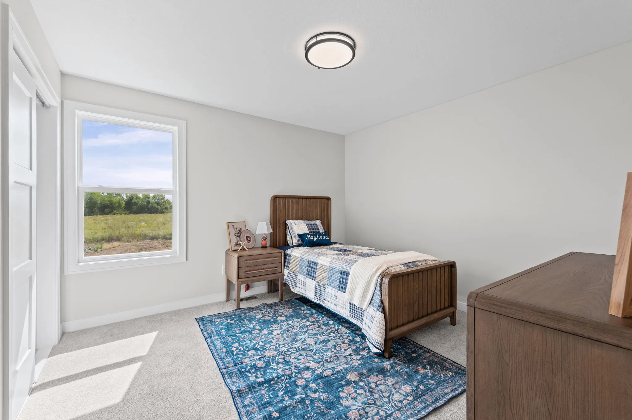 Bedroom with wood bed frame, blue patterned rug, nightstand, and large window overlooking green fields.