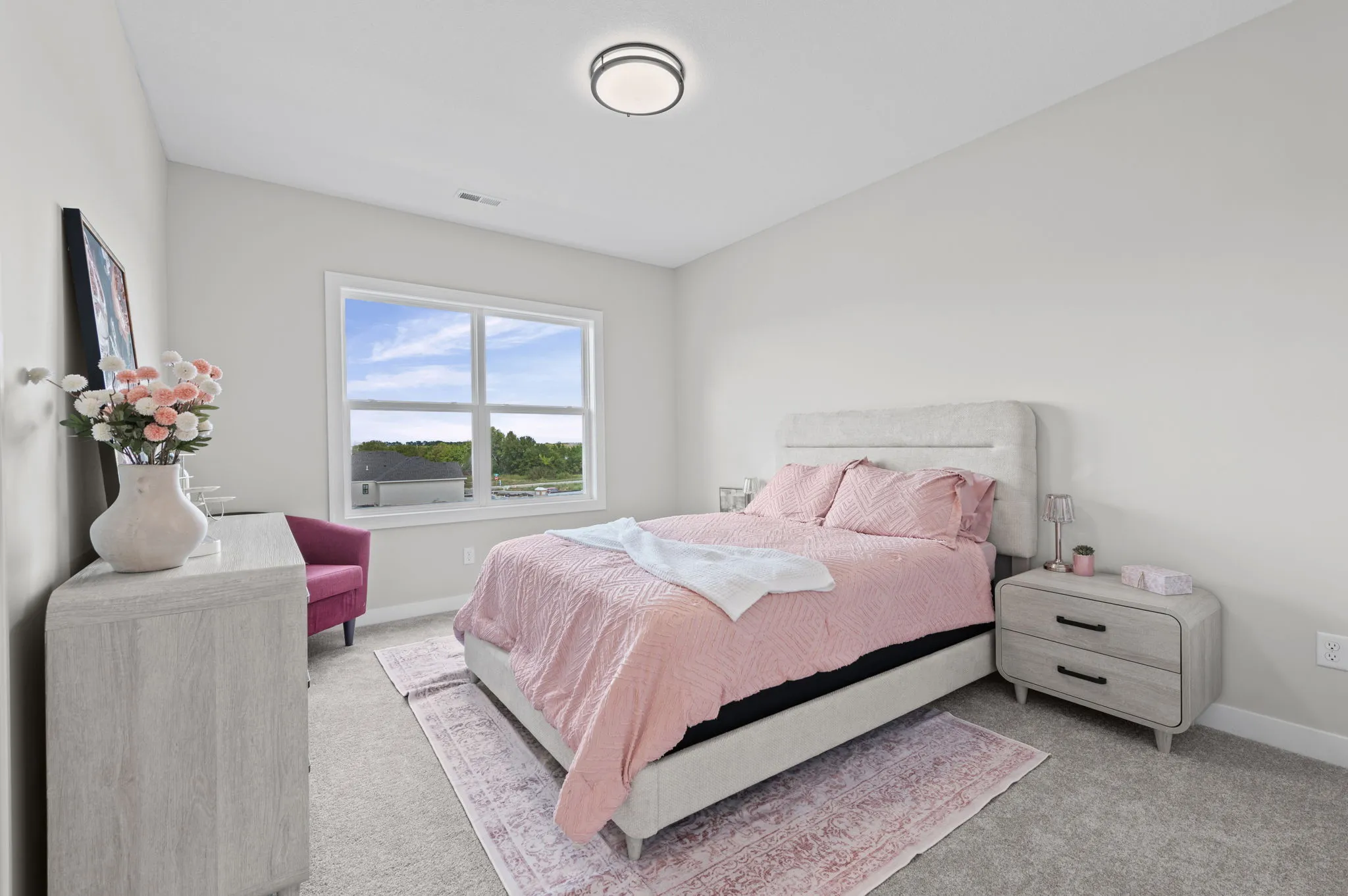 A bright bedroom with a bed dressed in pink bedding, nightstand, dresser with flowers and TV, and large windows overlooking
