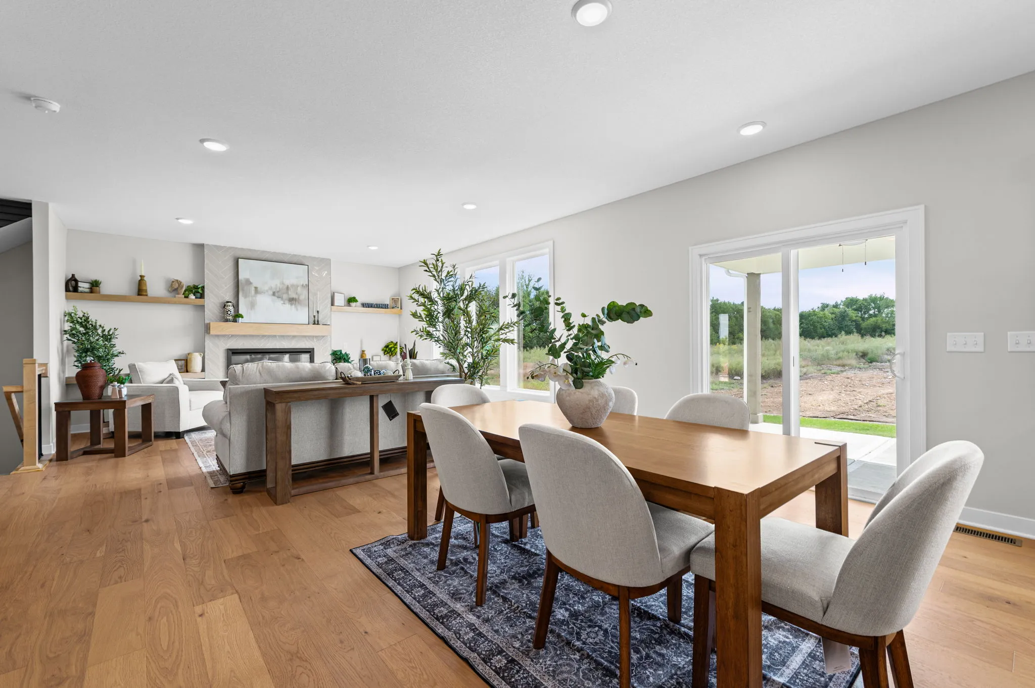 Open-concept living space with wood dining table, upholstered chairs, gray sofa, fireplace, and glass doors opening to
