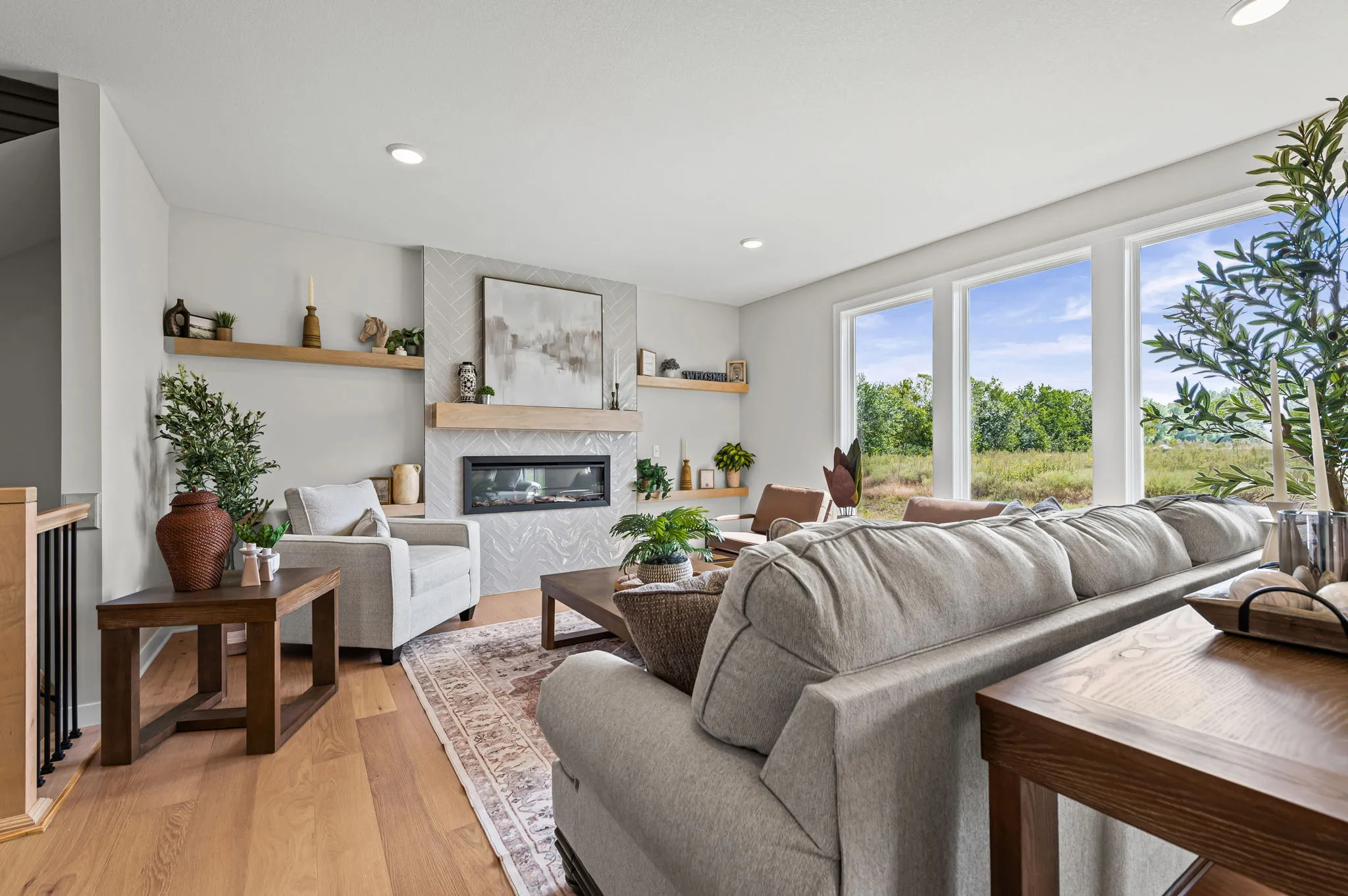 Living room with gray sofa, fireplace, wood furniture, and large windows overlooking trees.