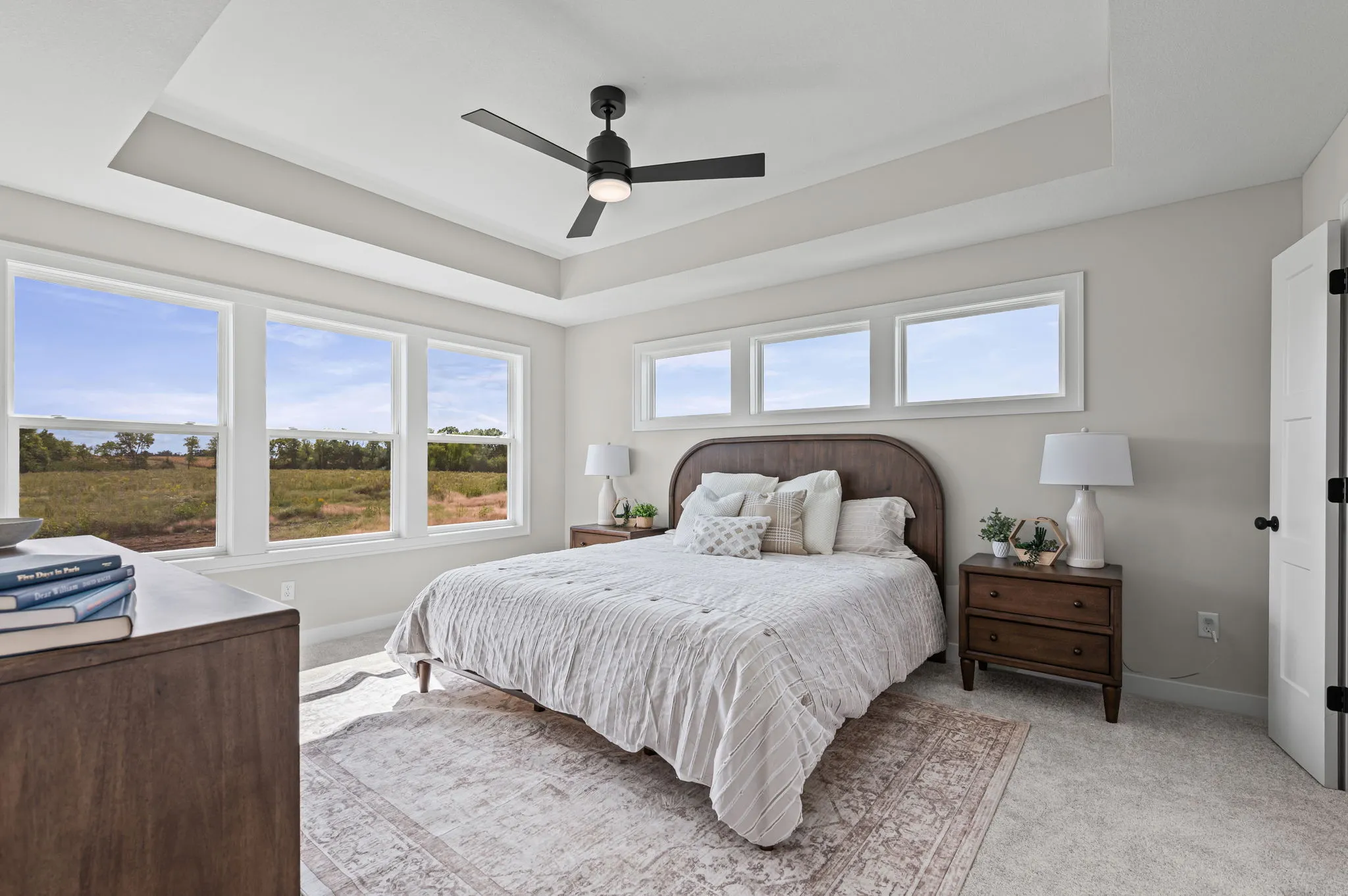 Master bedroom with tray ceiling, black ceiling fan, wood bed frame, nightstands with lamps, and large windows overlooking