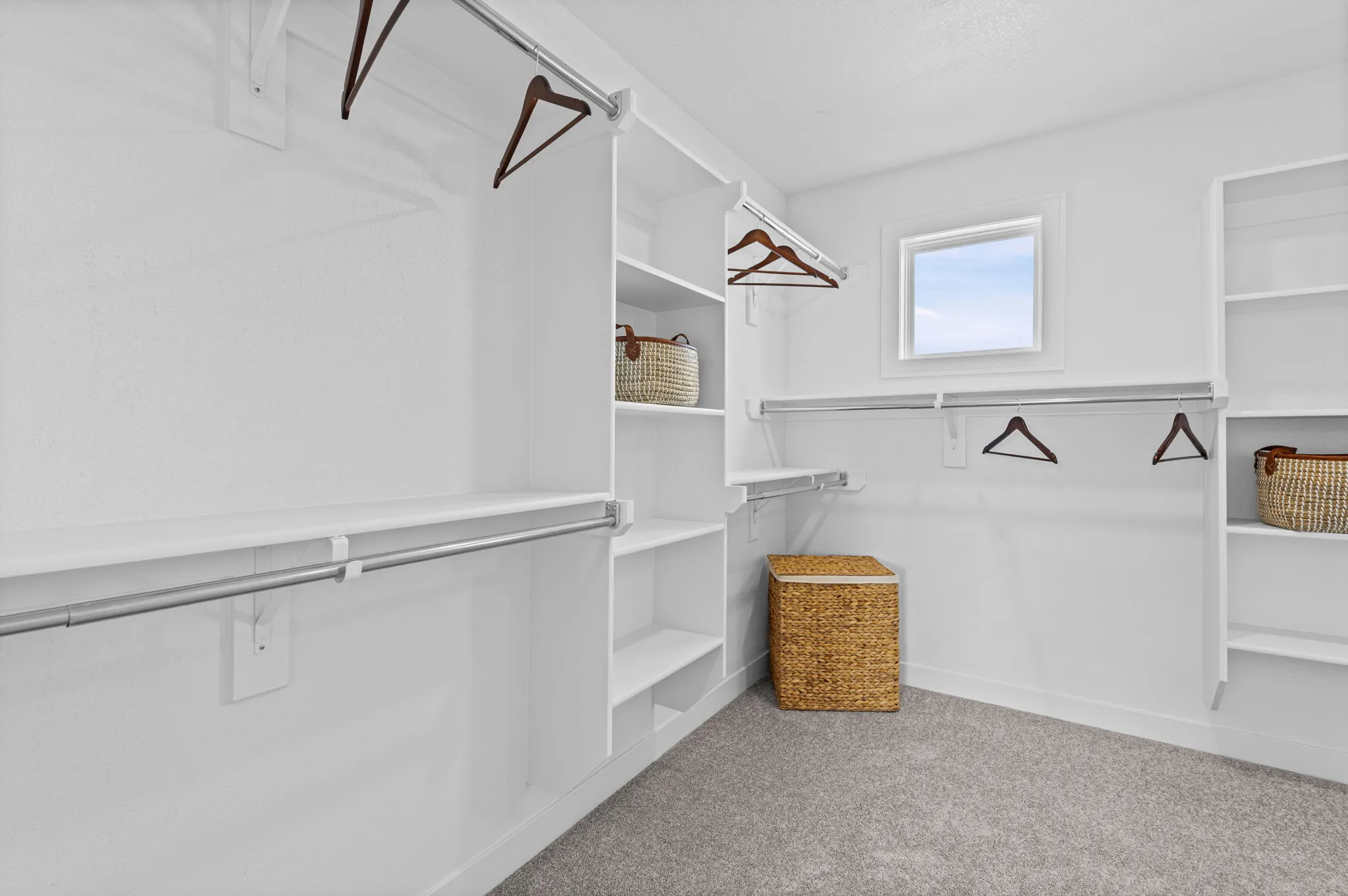 Walk-in closet with white shelving, hanging rods, wooden hangers, woven baskets, and a window.