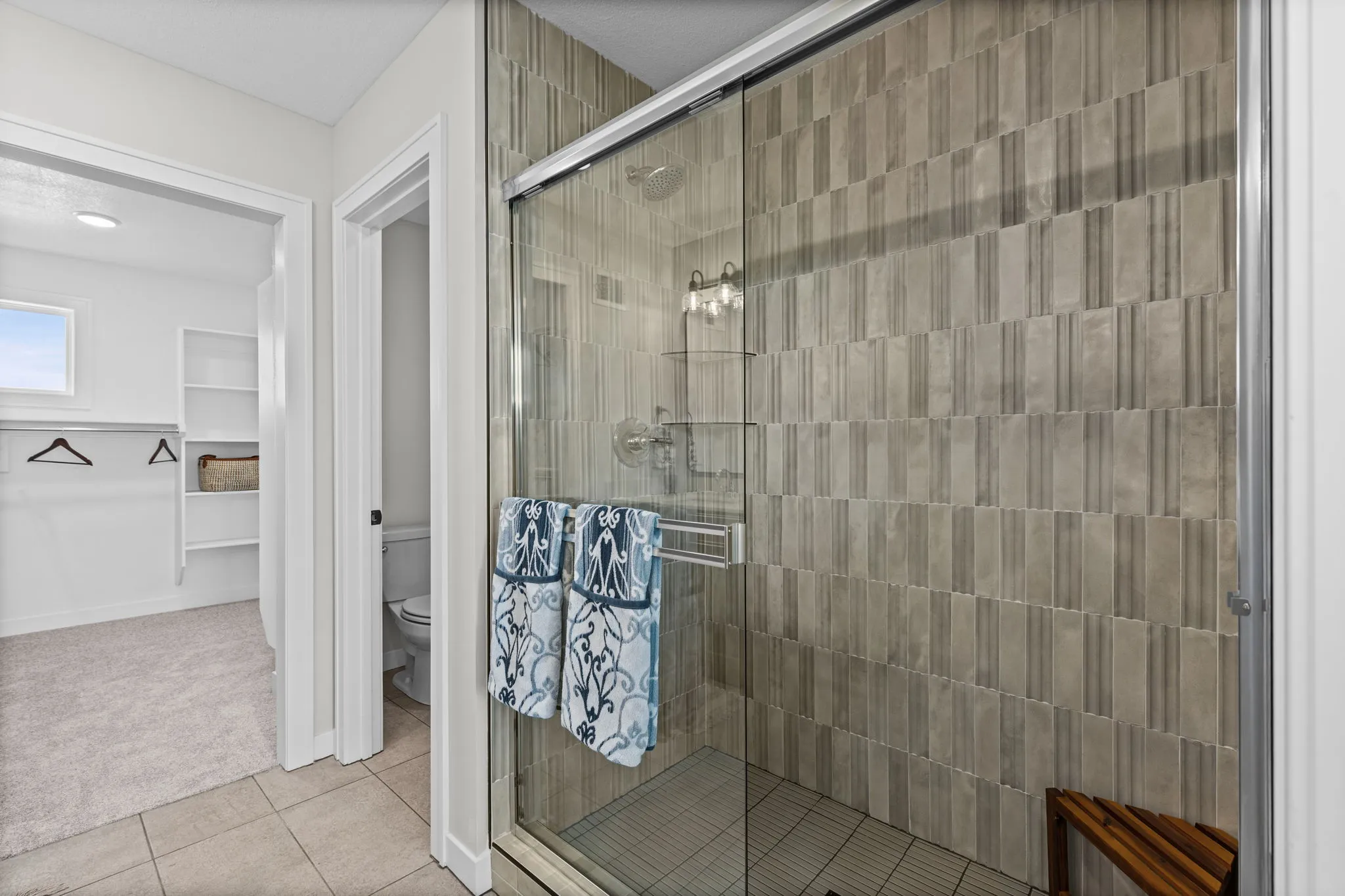 Walk-in shower with gray vertical tile walls, glass enclosure, towel bar, and showerhead, adjoining bathroom with toilet and