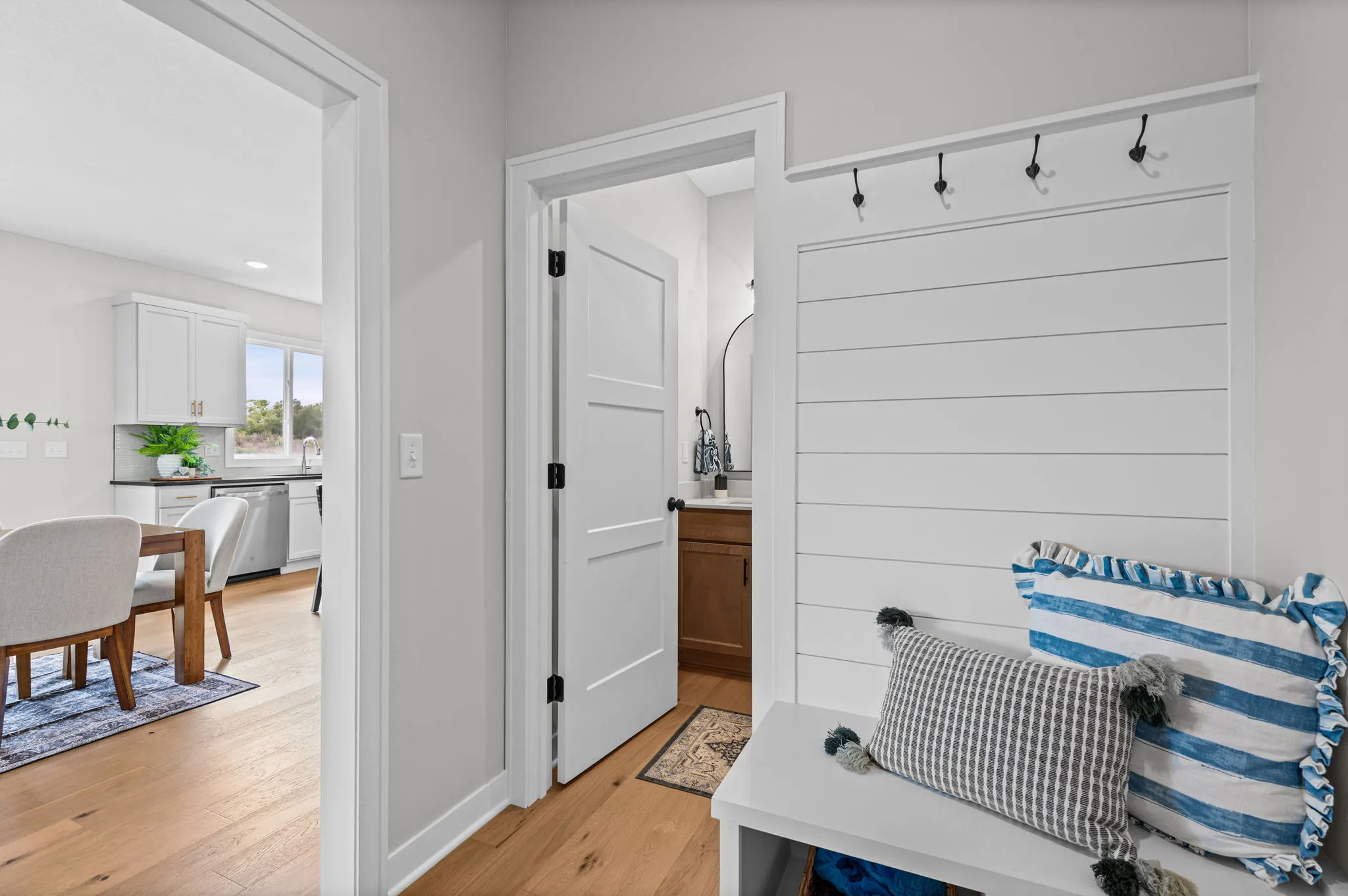 Mudroom with white shiplap walls, white paneled doors, coat hooks, bench with striped pillows, and view into kitchen with