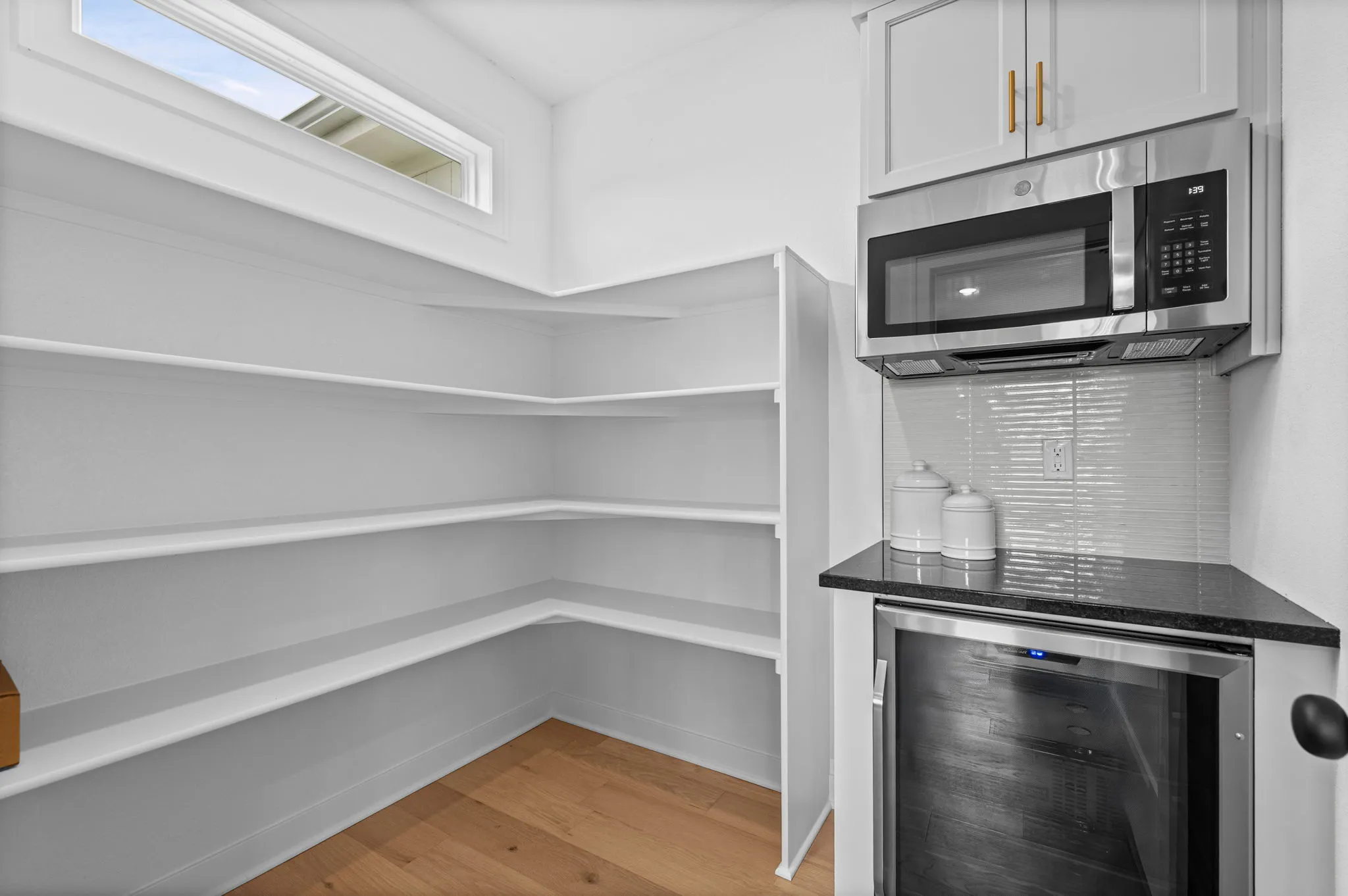 Walk-in pantry with empty white shelving and wood flooring adjacent to stainless steel microwave and oven.