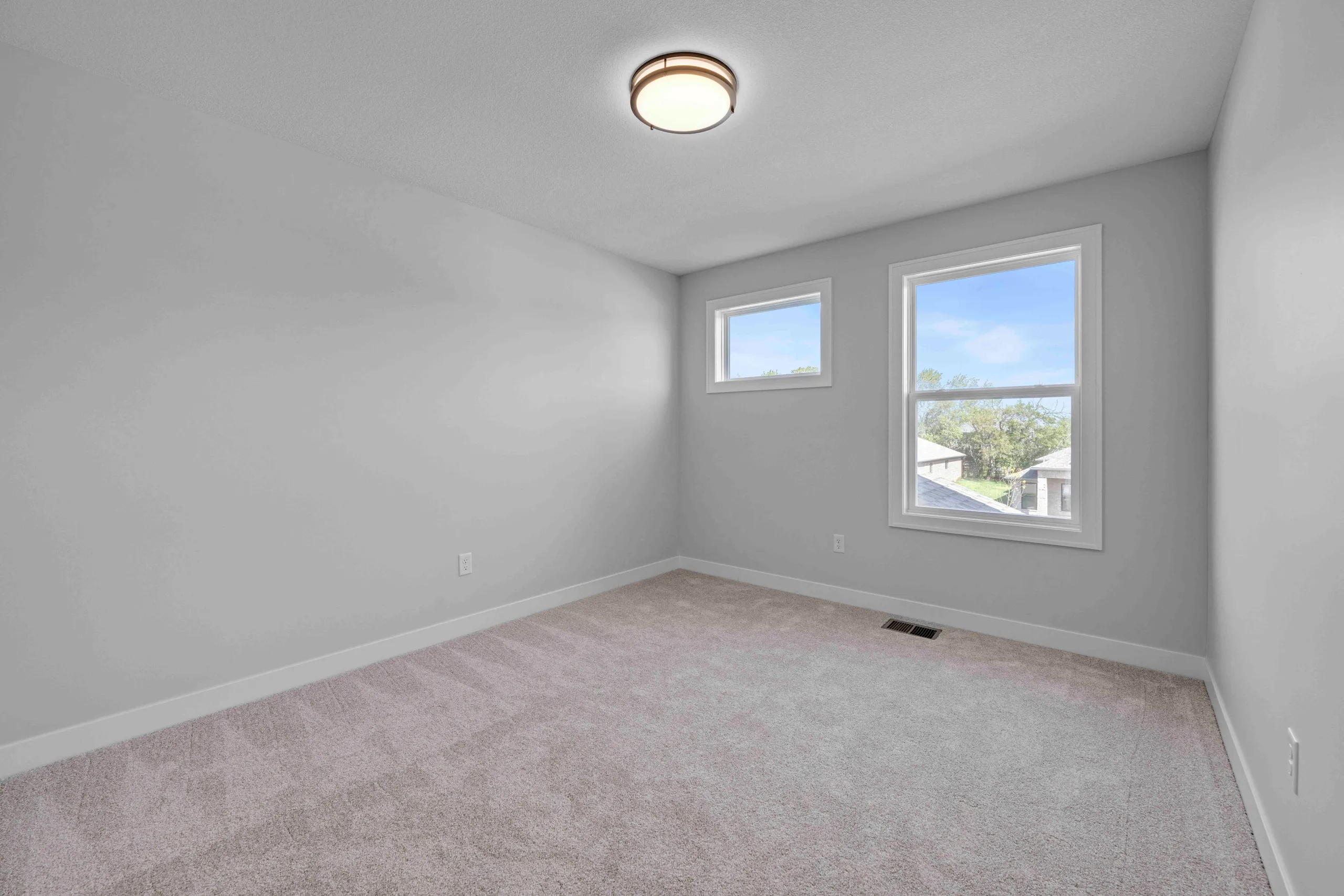 Empty bedroom with gray walls, beige carpet, recessed ceiling light, and two windows with views of trees.