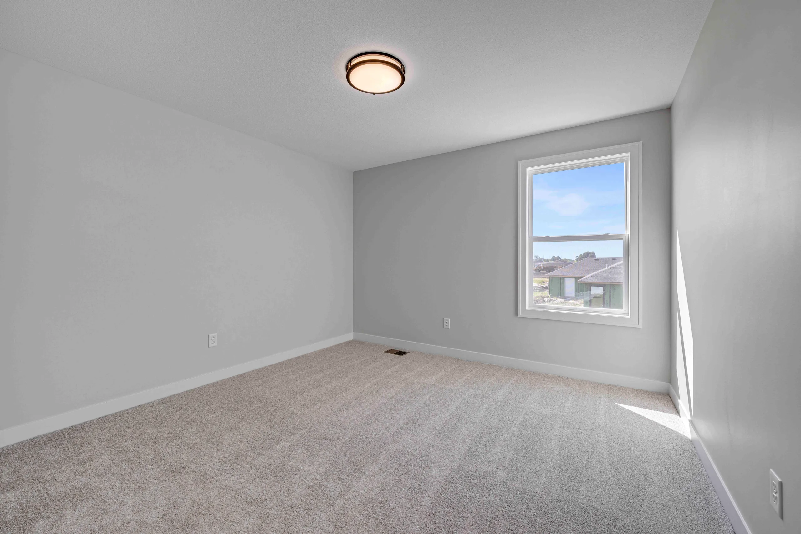 Empty bedroom with gray walls, beige carpet, ceiling light fixture, and window overlooking neighborhood homes.