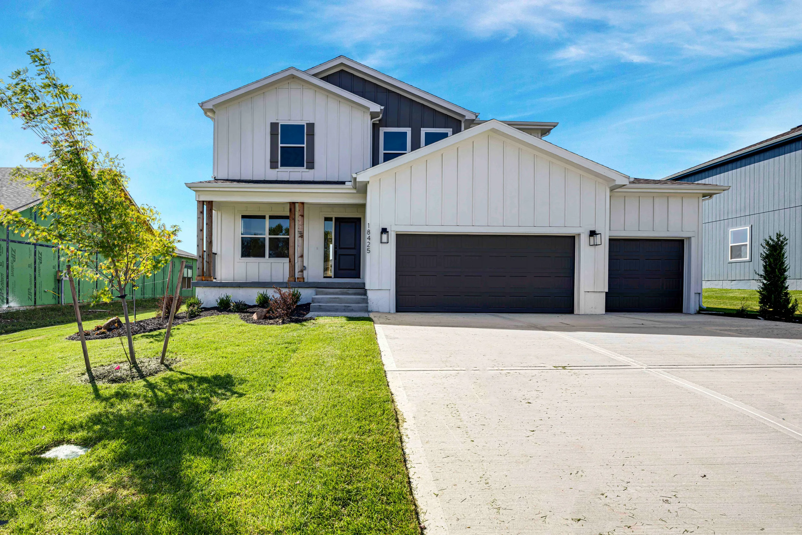 Two-story home with white vertical siding, black garage doors, front porch, and landscaped lawn with young trees.