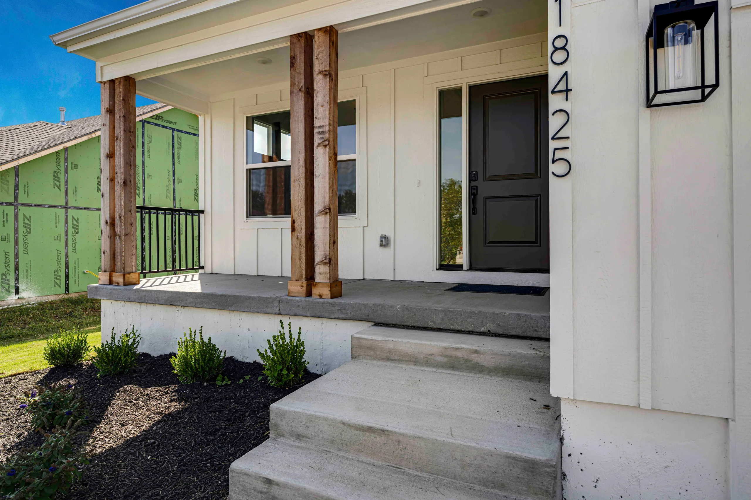 Front porch with concrete steps, black front door, wooden support columns, and landscaping bed below.