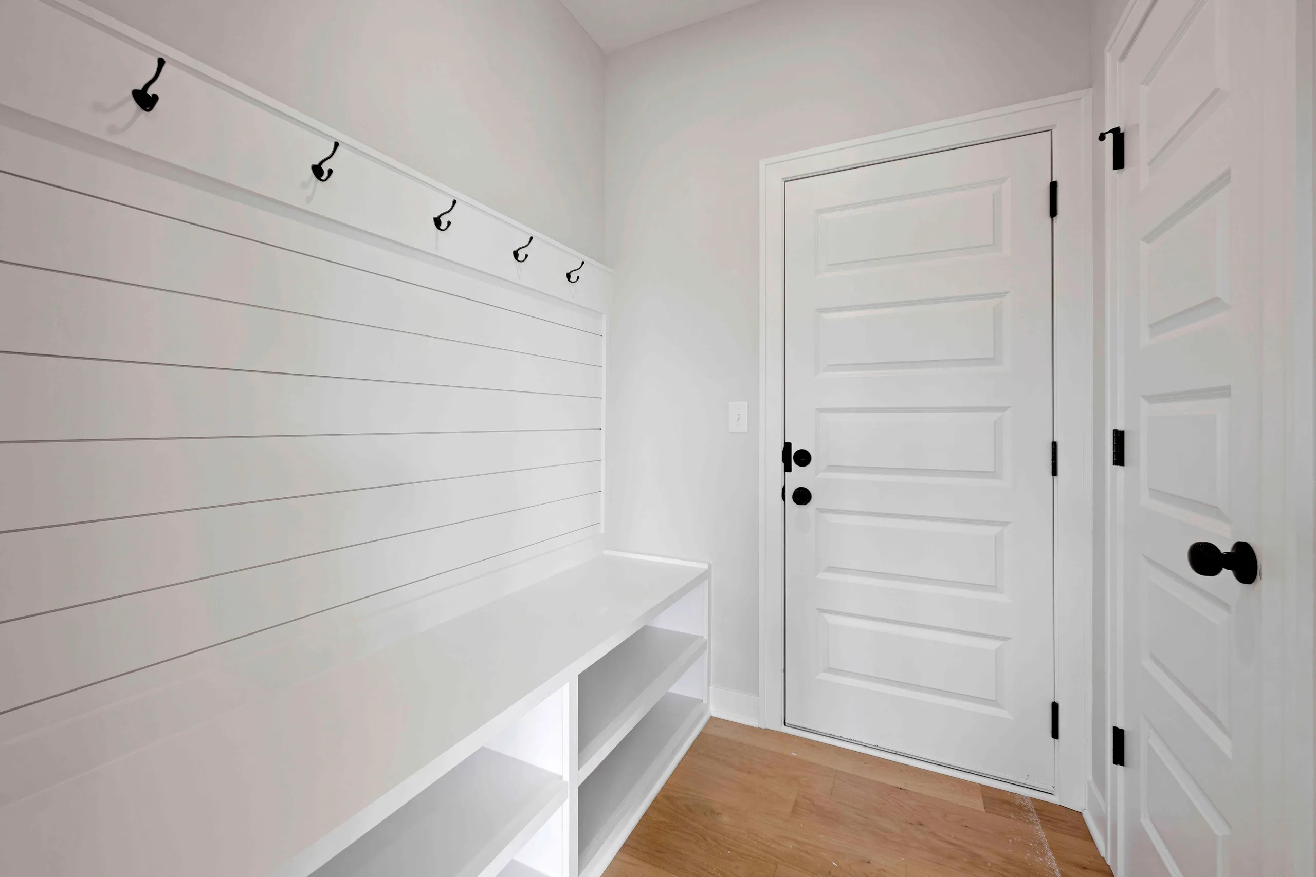 White mudroom with built-in shelving, black hooks, white panel doors with black hardware, and wood flooring.