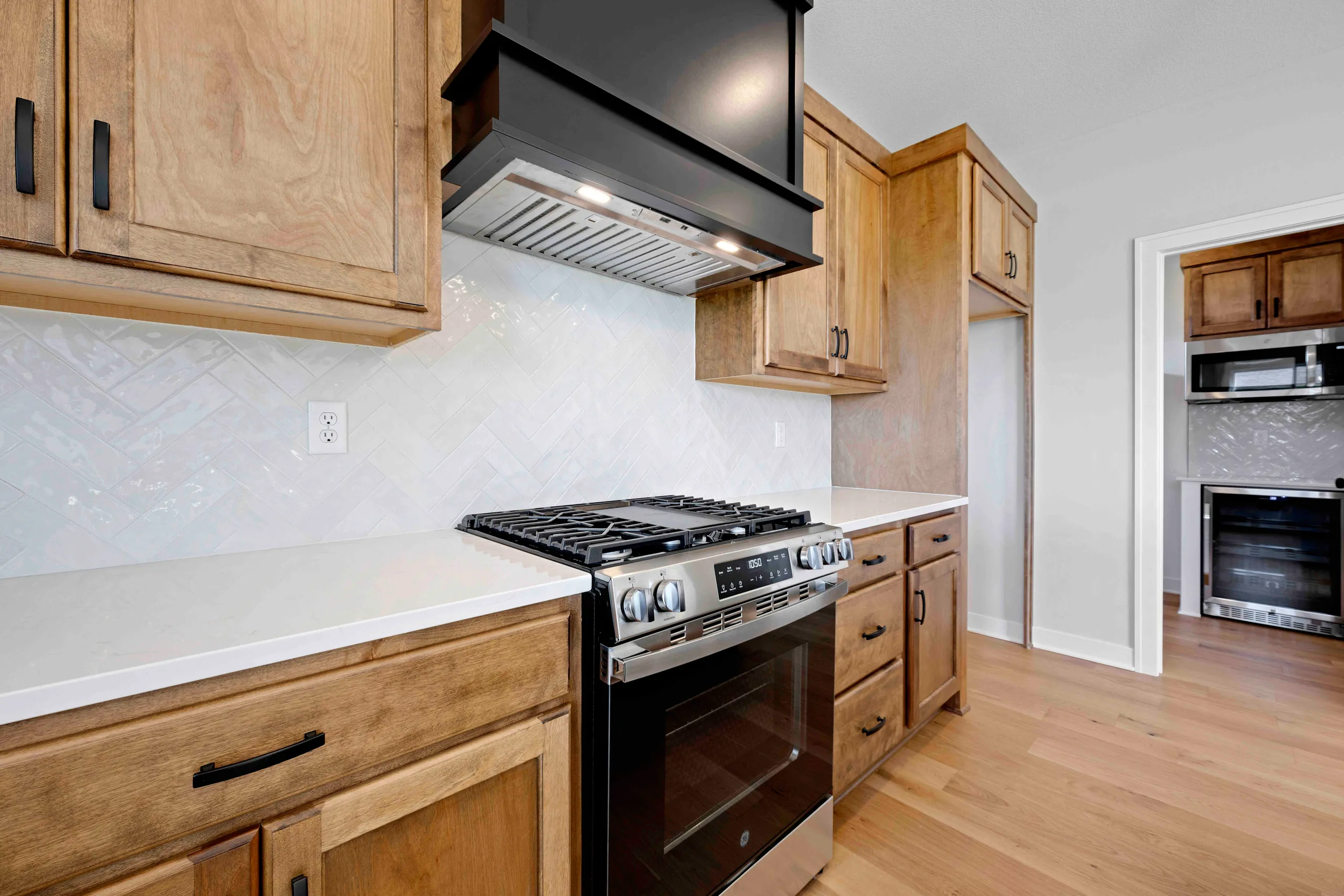 Kitchen with wooden cabinetry, white countertops, stainless steel gas range, black range hood, and herringbone tile