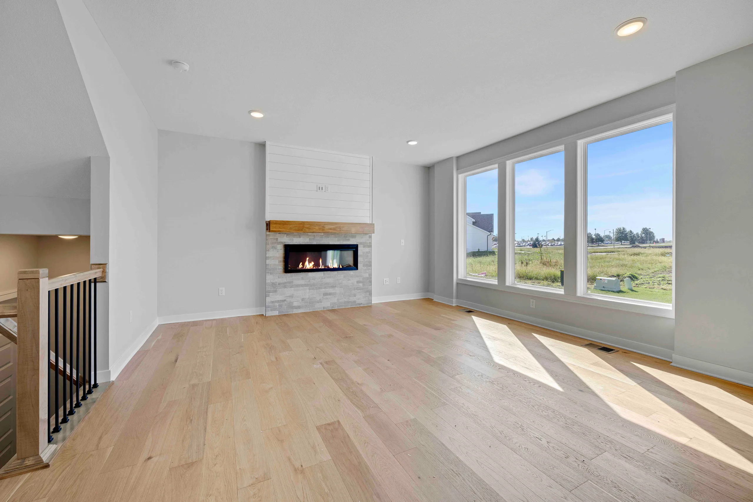 Open living room with fireplace, wood flooring, large windows overlooking grassland, and staircase with black railing.