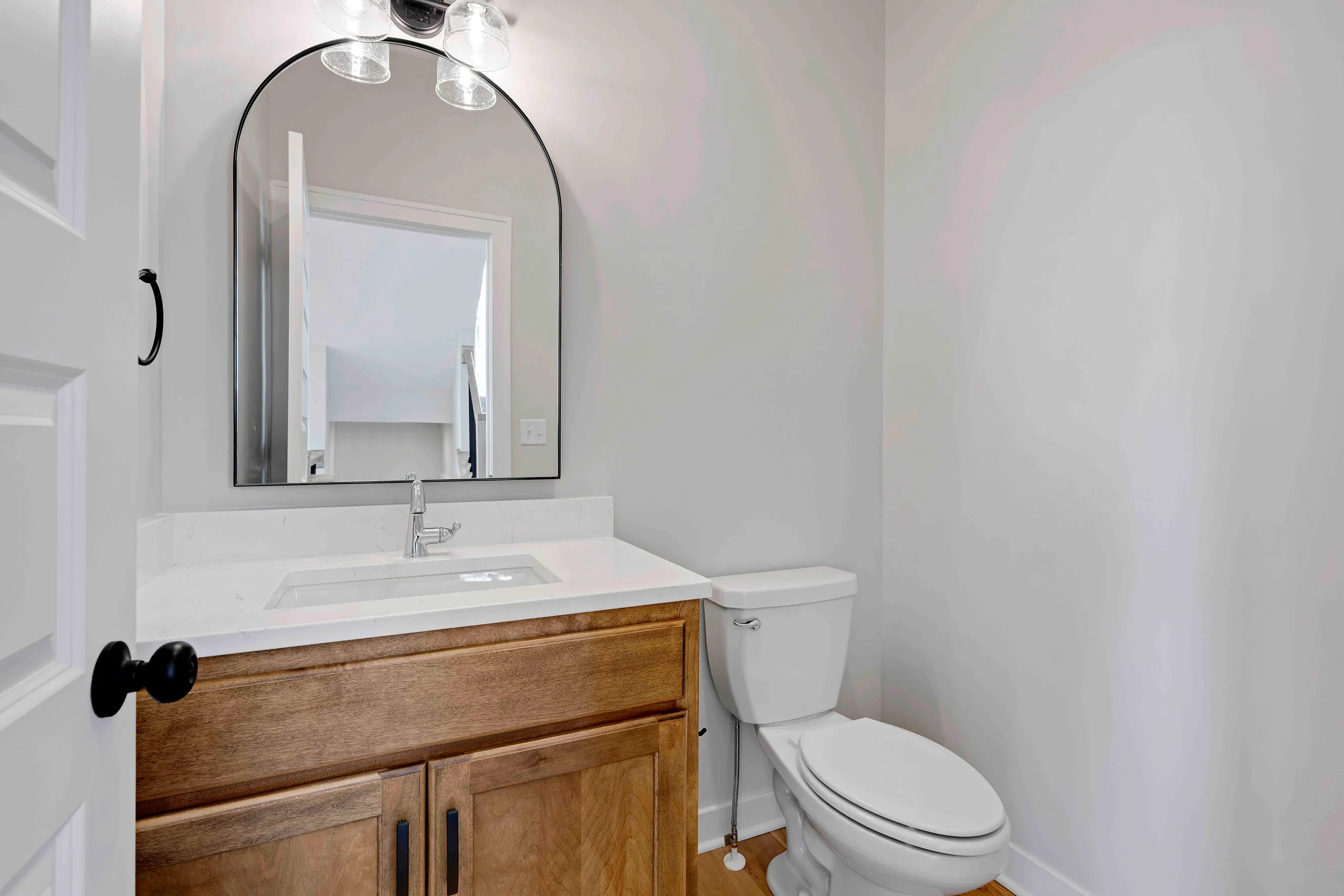 Bathroom with white walls, arched mirror with black frame above wooden vanity, white countertop, single sink, and white