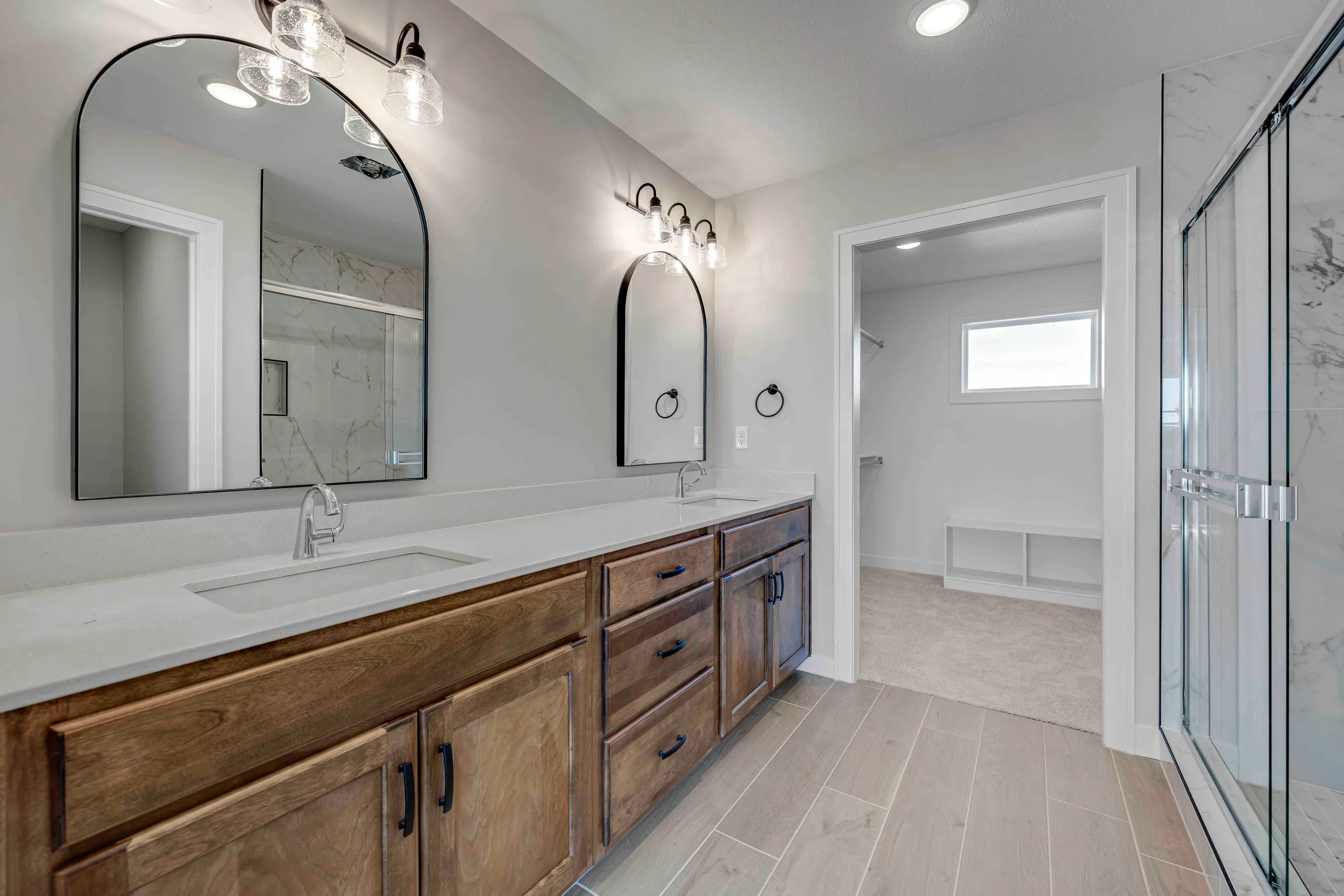 Bathroom with wood vanity, white countertop, dual arched mirrors, and glass shower enclosure on the right.