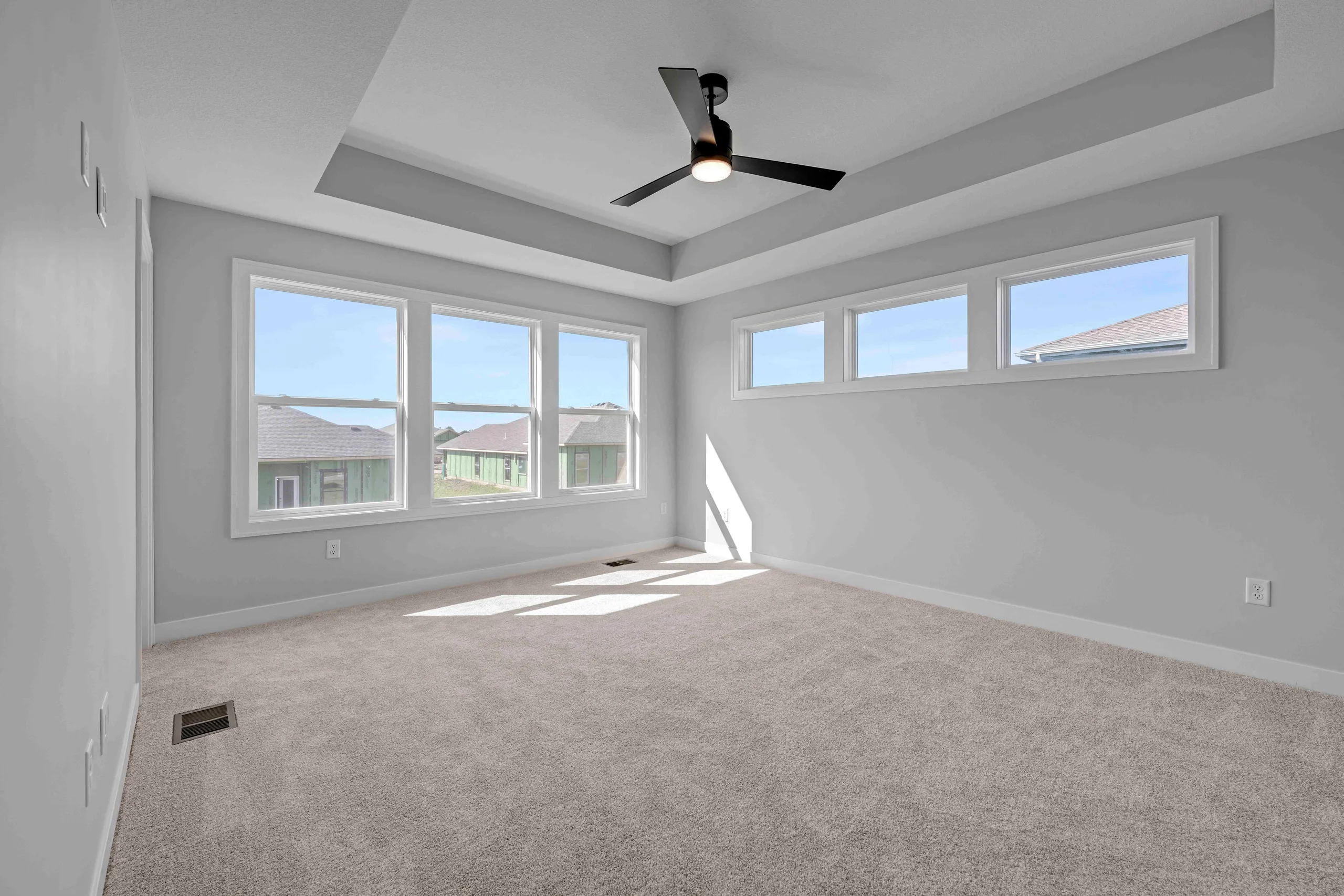 Bedroom with gray carpet, white walls, ceiling fan, and large windows overlooking neighboring homes.