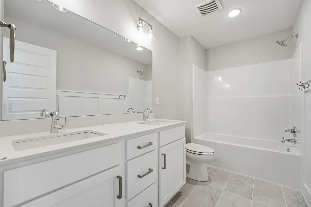 White bathroom with double sink vanity, large mirror with lighting, toilet, and tub-shower combination.