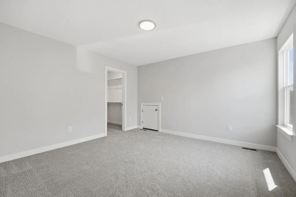 Empty bedroom with gray carpet, white walls, recessed lighting, window, closet, and small utility door.