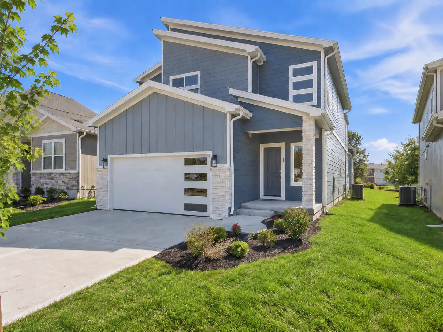 Two-story home with gray and white siding, white garage door, front entry, landscaping, and neighboring houses on a sunny