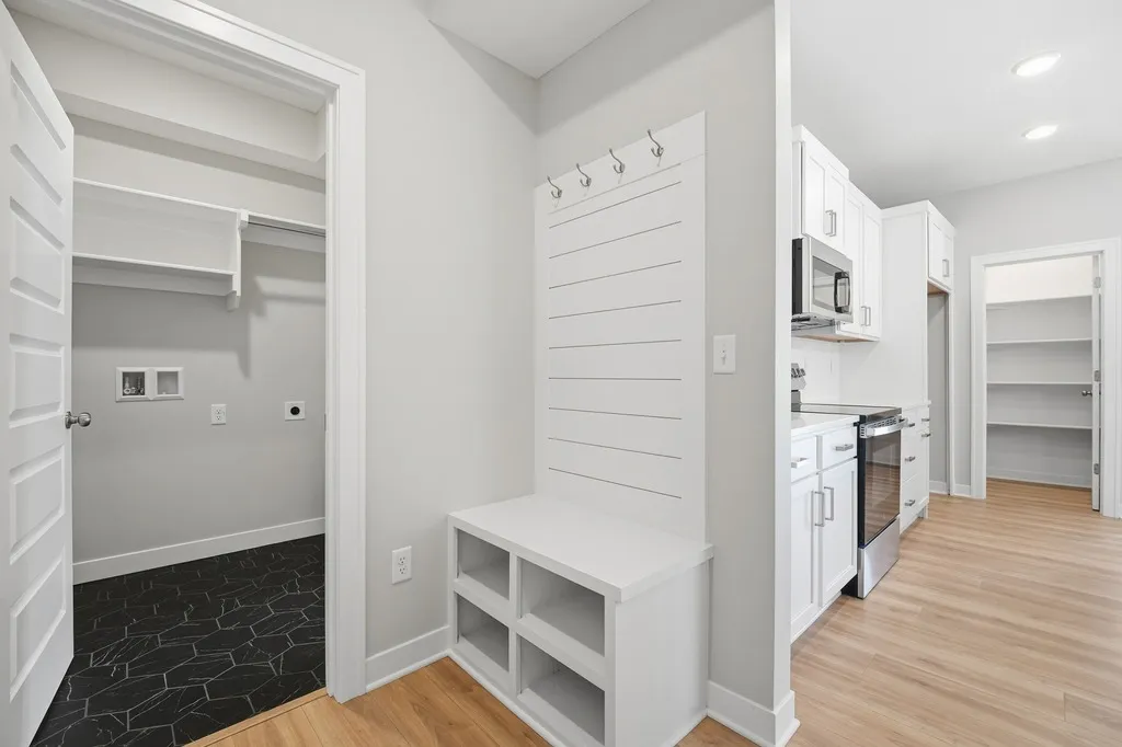 Mudroom with white built-in bench and shelving, wall hooks, and doorway to laundry room with black tile flooring.