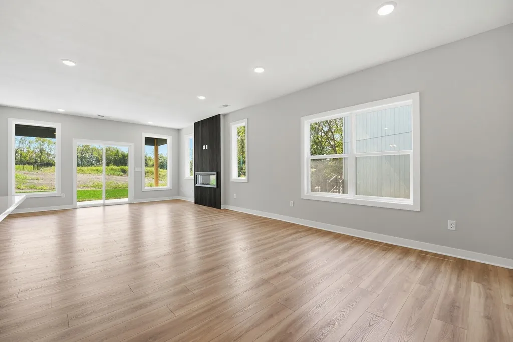 Empty living room with wood flooring, gray walls, recessed lighting, black fireplace, and large windows overlooking green