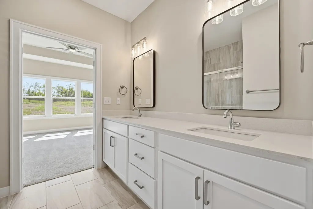 Bathroom with dual sinks, white vanity, framed mirrors, and door leading to bedroom with outdoor view.