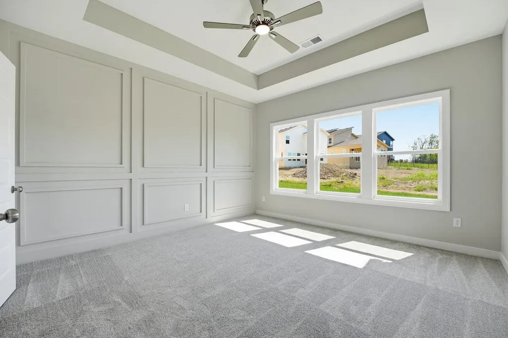Empty room with gray carpet, white paneled walls, tray ceiling with ceiling fan, and large windows overlooking neighborhood.