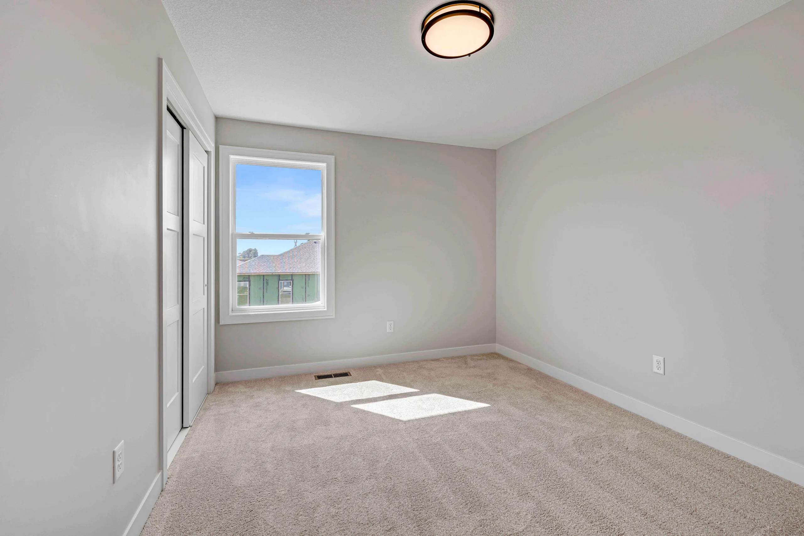 Empty bedroom with beige carpeting, white walls, window with view of neighboring homes, and ceiling light fixture.
