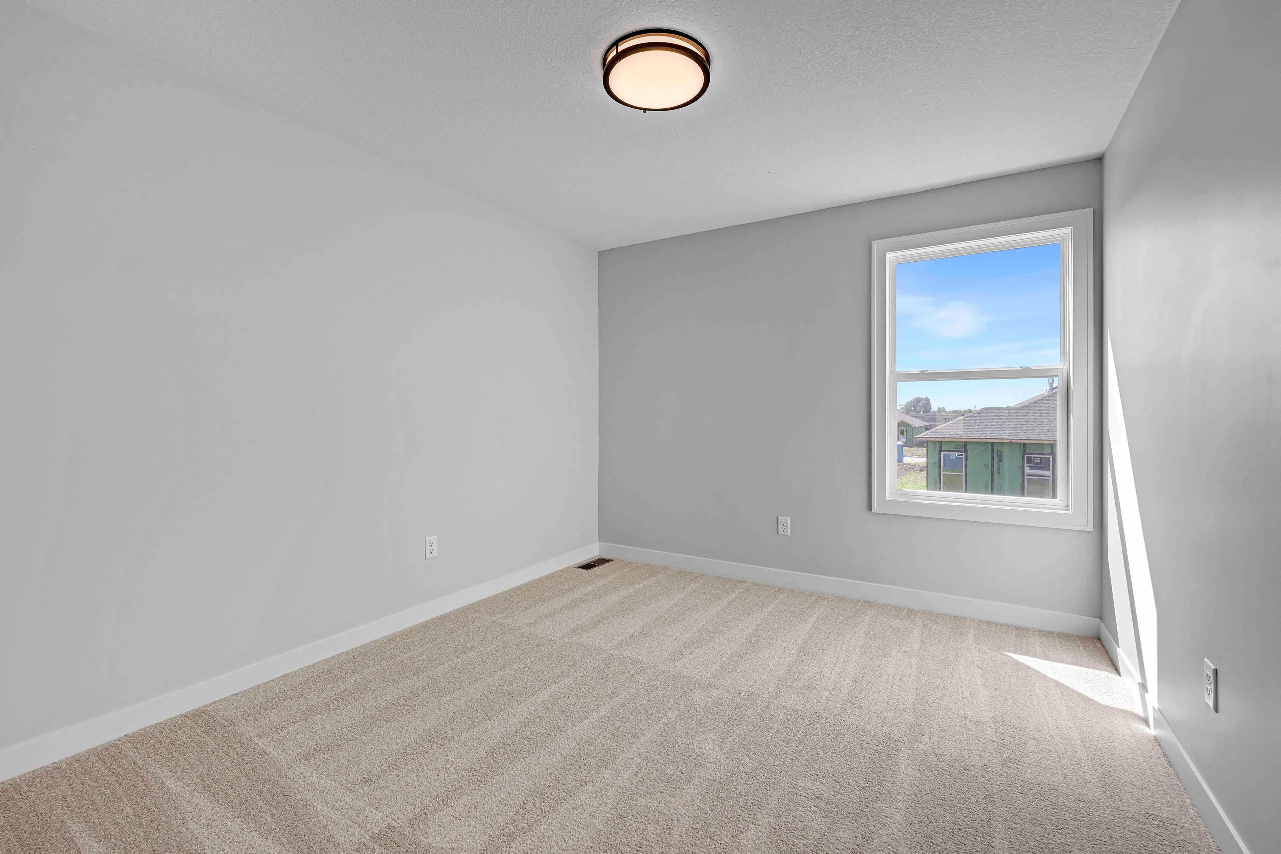 Empty bedroom with gray walls, beige carpet, ceiling light fixture, and window overlooking neighborhood.
