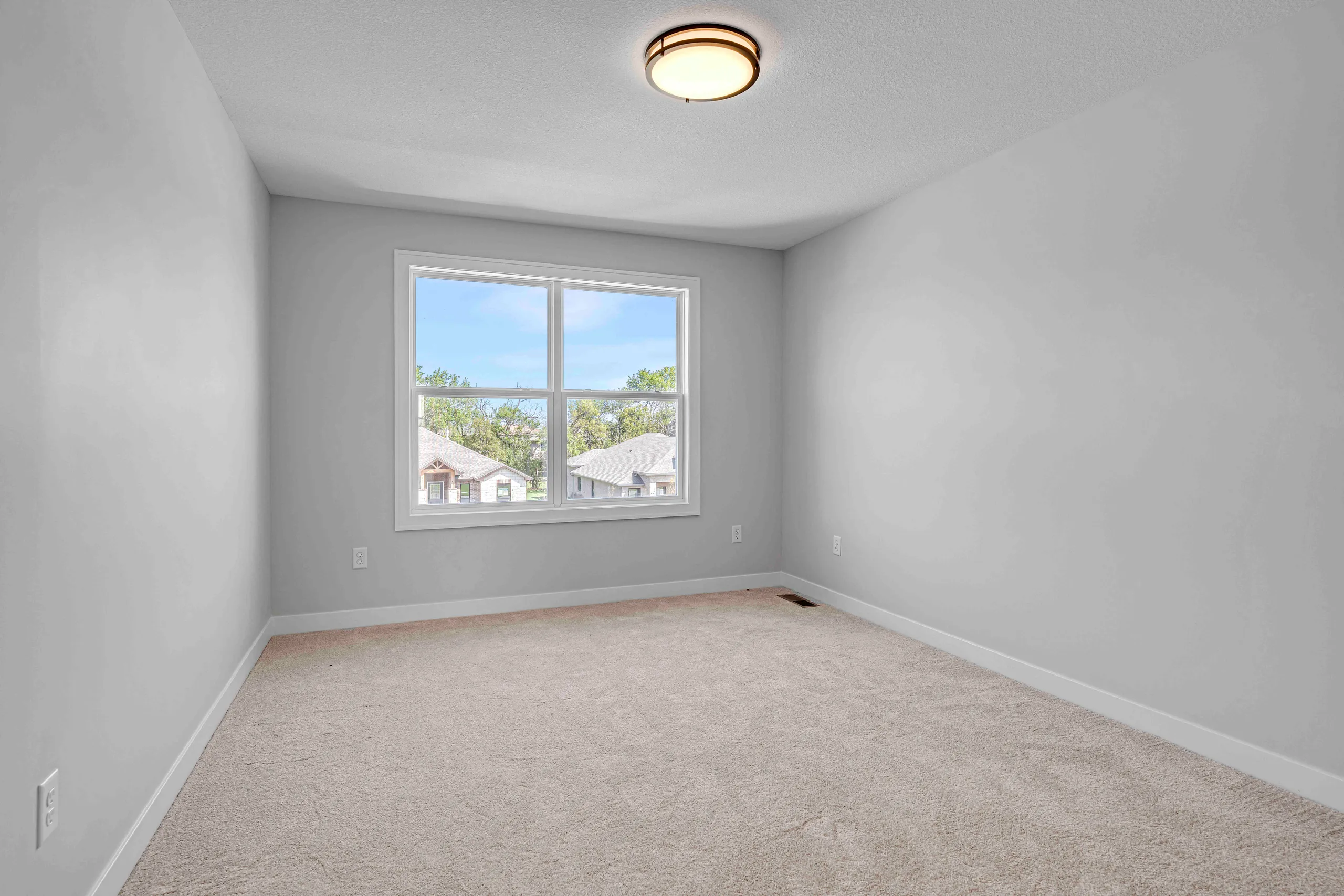 Empty bedroom with gray walls, beige carpet, ceiling light fixture, and multi-pane window overlooking neighborhood homes.