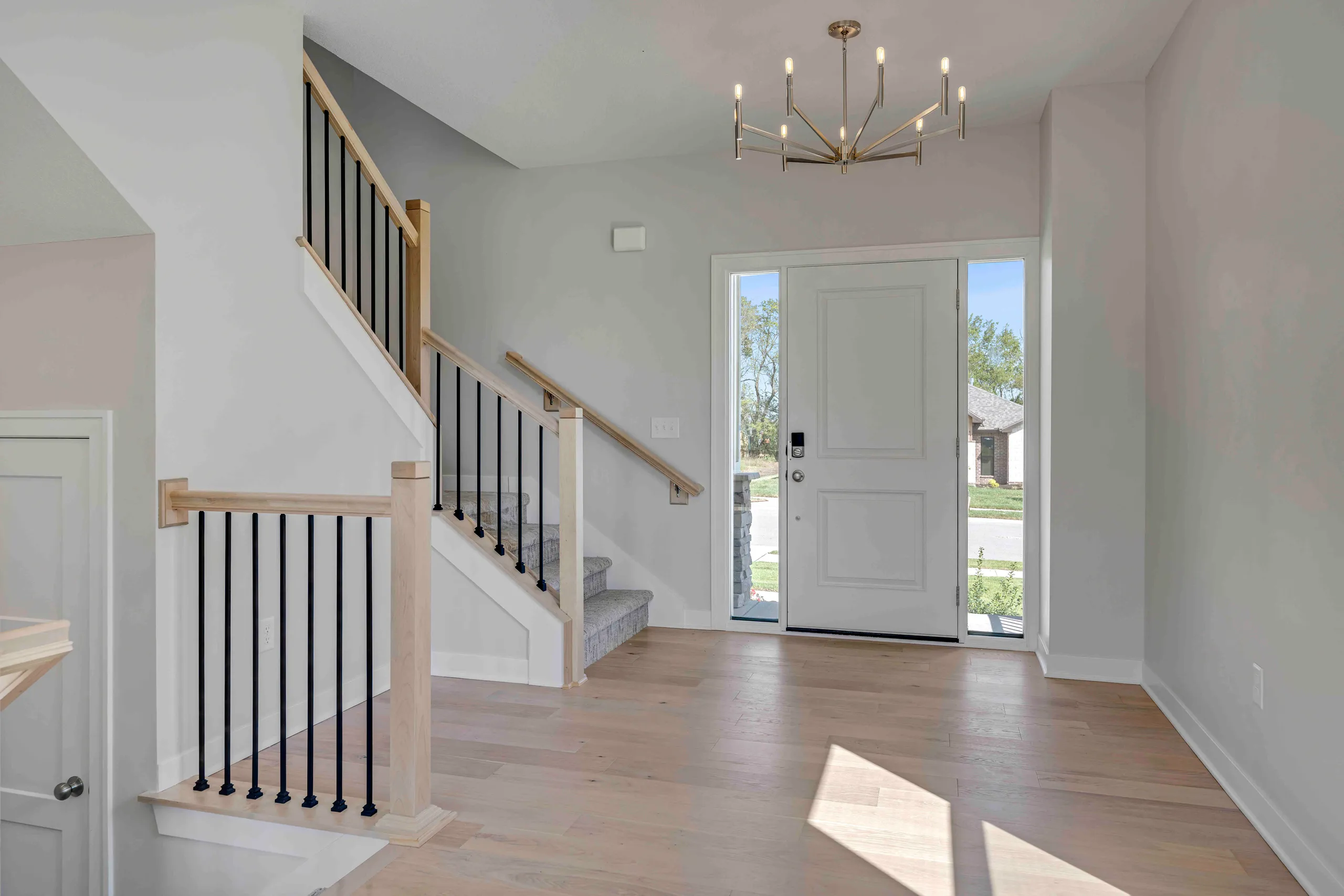 Foyer with wooden staircase, black metal railings, brass chandelier, and white front door with sidelights.