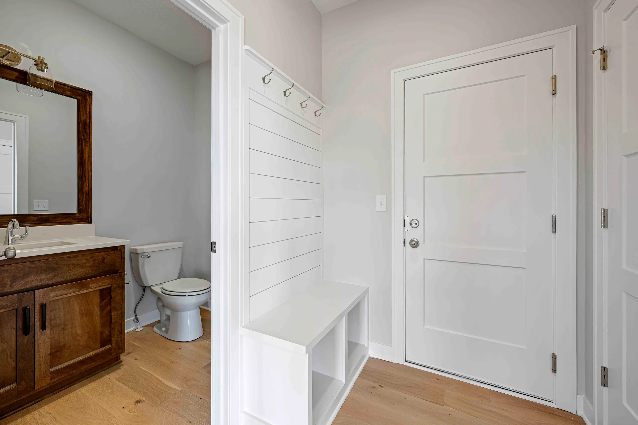 Bathroom and mudroom with wood vanity, toilet, mirror, white shiplap wall, built-in bench, and white door.