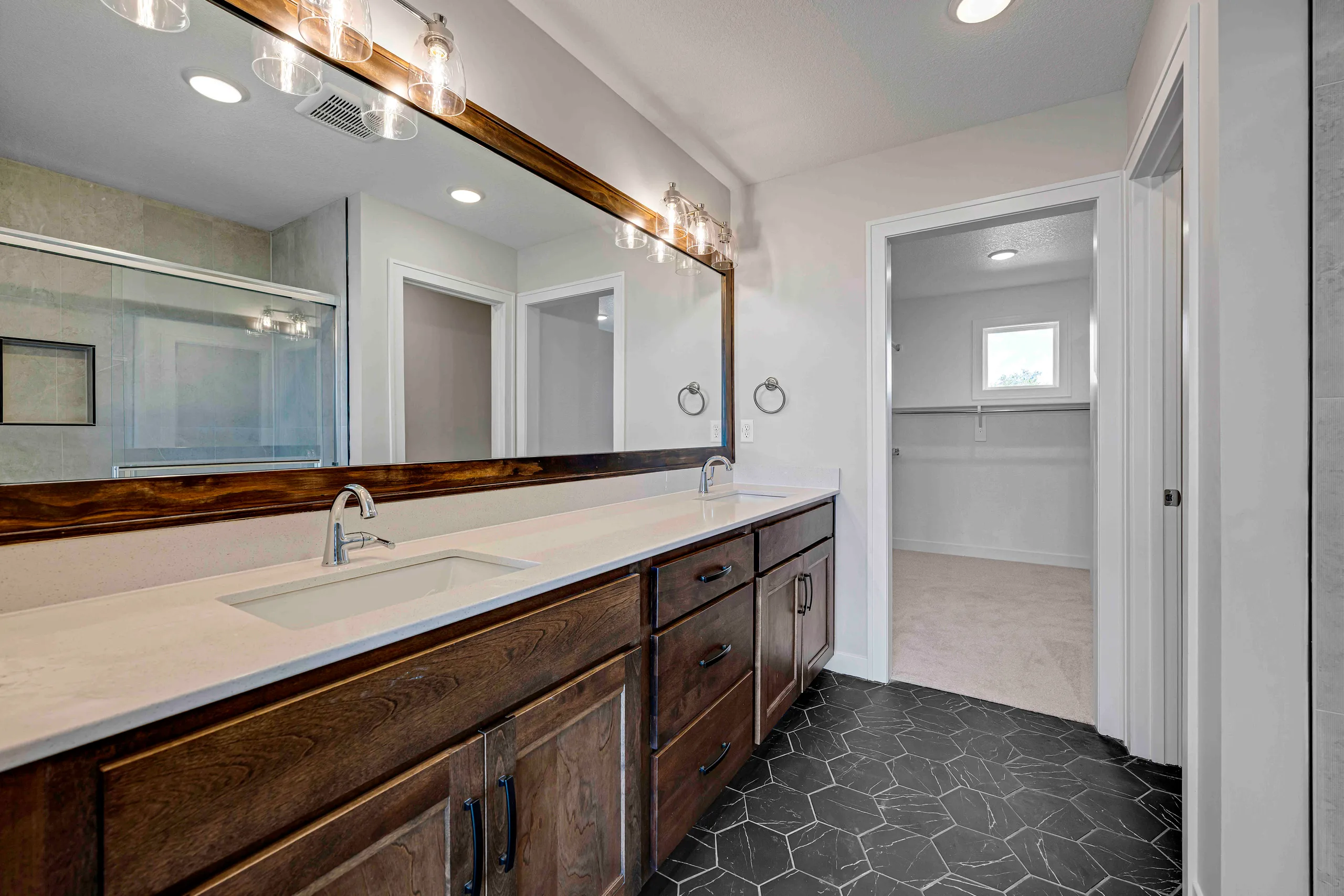 Master bathroom with dark wood double vanity, white countertop, large mirror, and black hexagonal tile flooring.