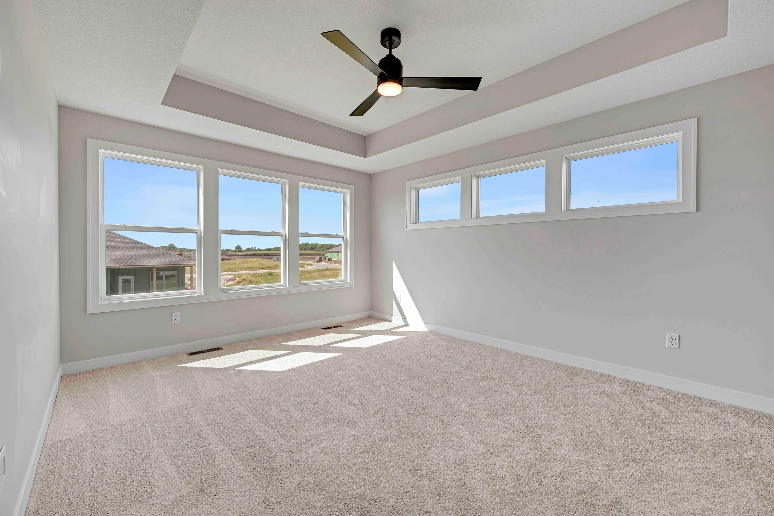 Empty room with beige carpet, white walls, tray ceiling with ceiling fan, and multiple windows overlooking open landscape.