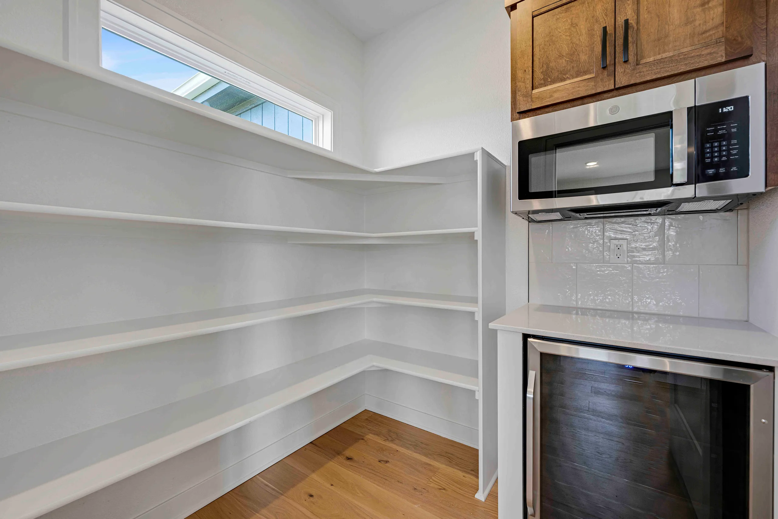 Pantry with white shelving, wood flooring, and a window, adjoining kitchen with stainless steel appliances and white subway