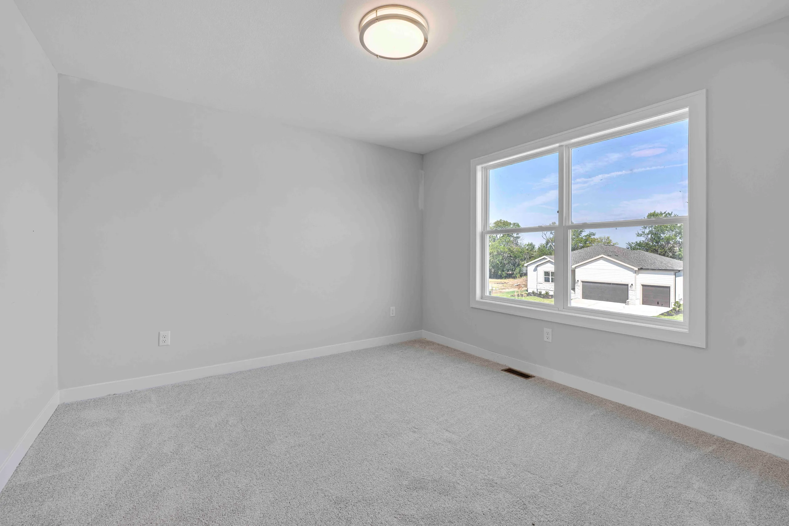 Empty bedroom with gray carpet, white walls, ceiling light fixture, and large window overlooking neighboring homes.
