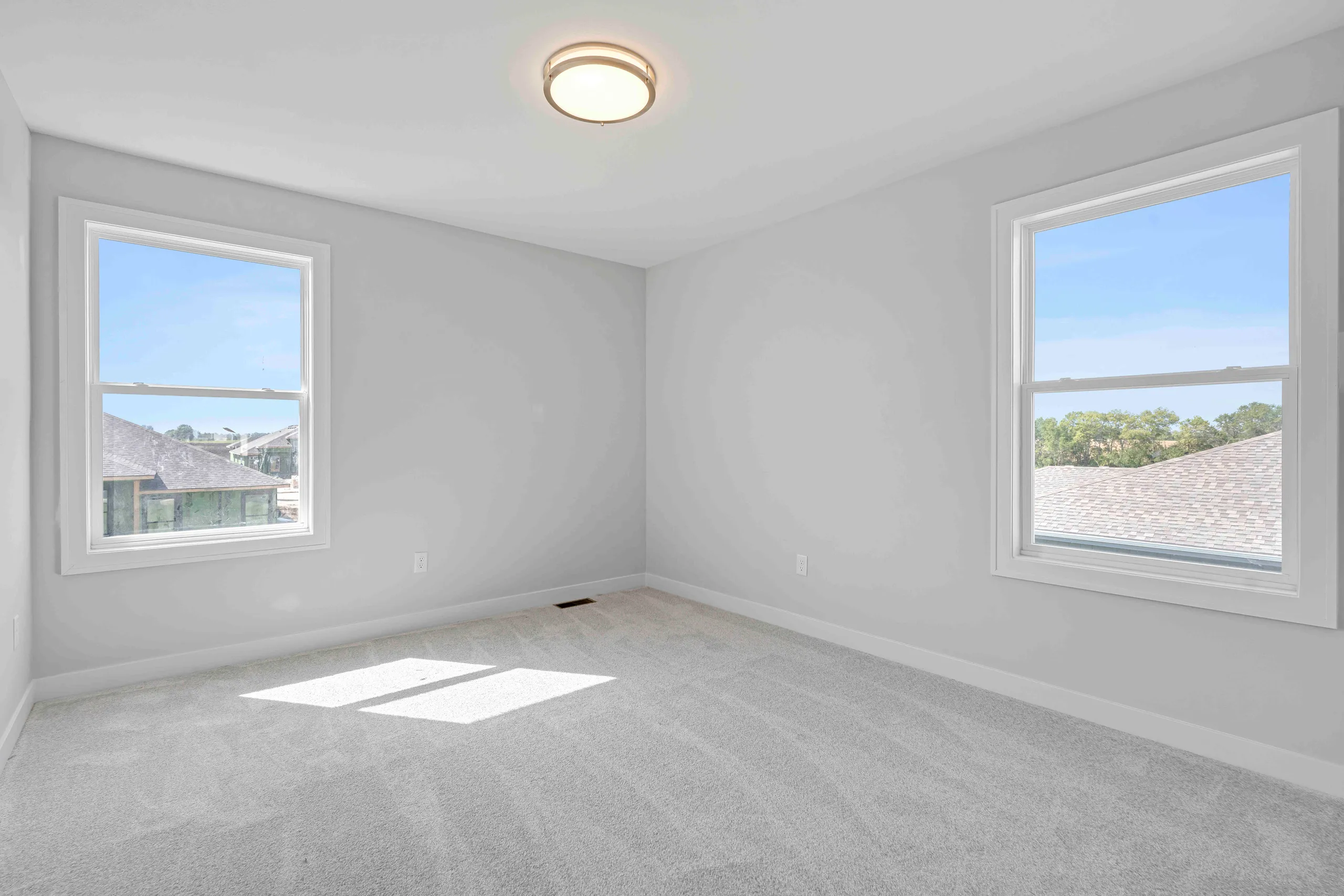 Empty bedroom with gray carpet, white walls, two windows overlooking neighborhood, and ceiling light fixture.