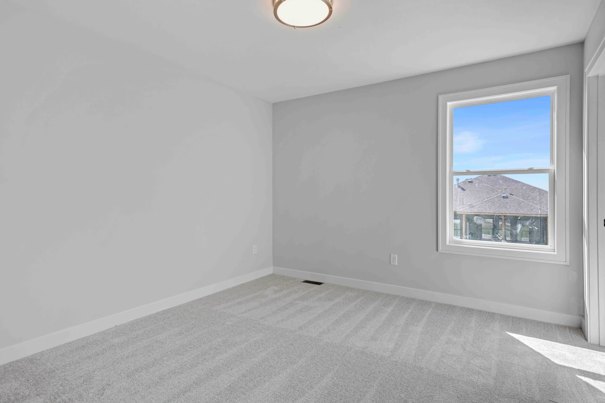 Empty bedroom with gray carpet, gray walls, ceiling light fixture, and window overlooking residential rooflines.