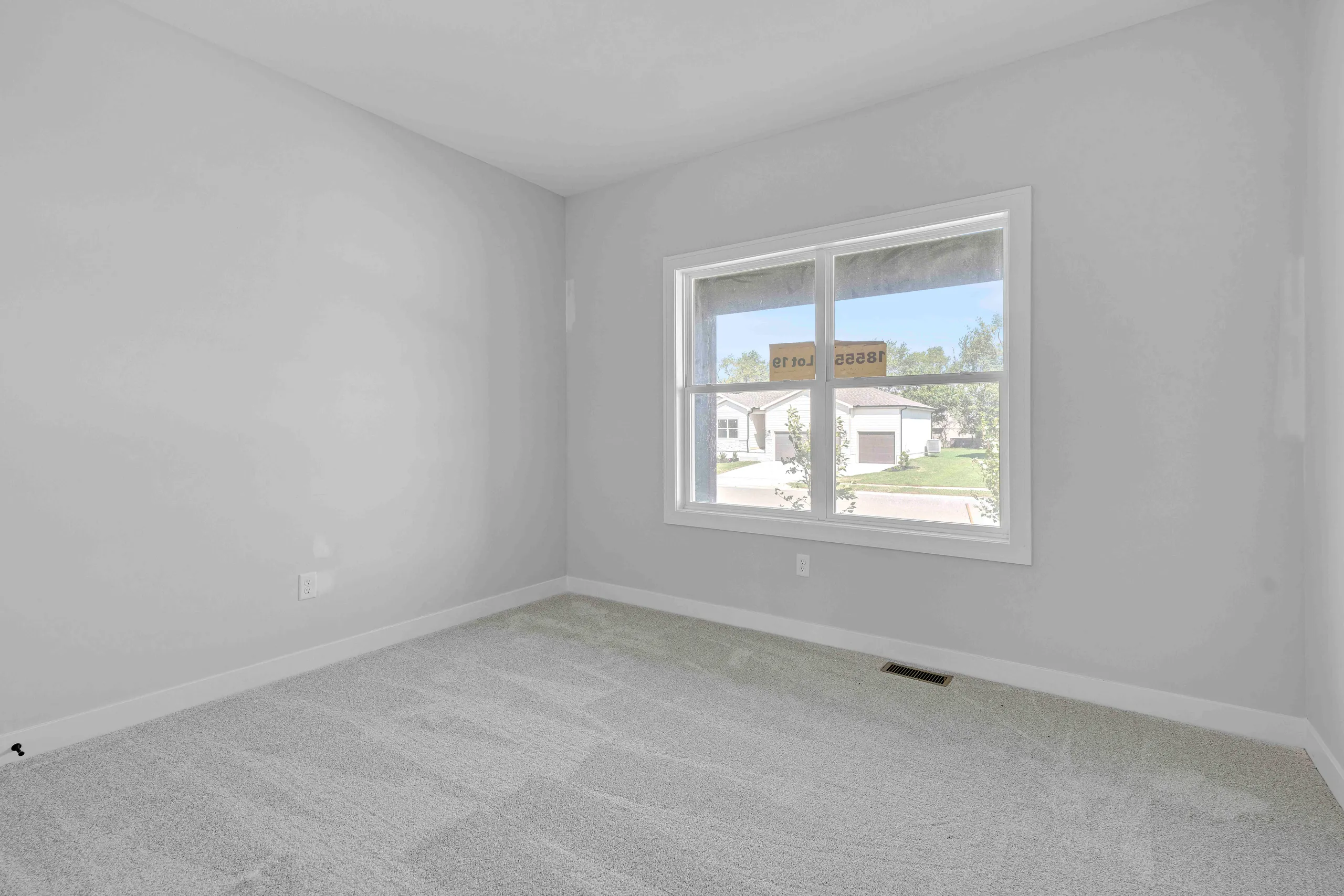 Empty bedroom with gray carpet, white walls, and large window overlooking residential neighborhood.