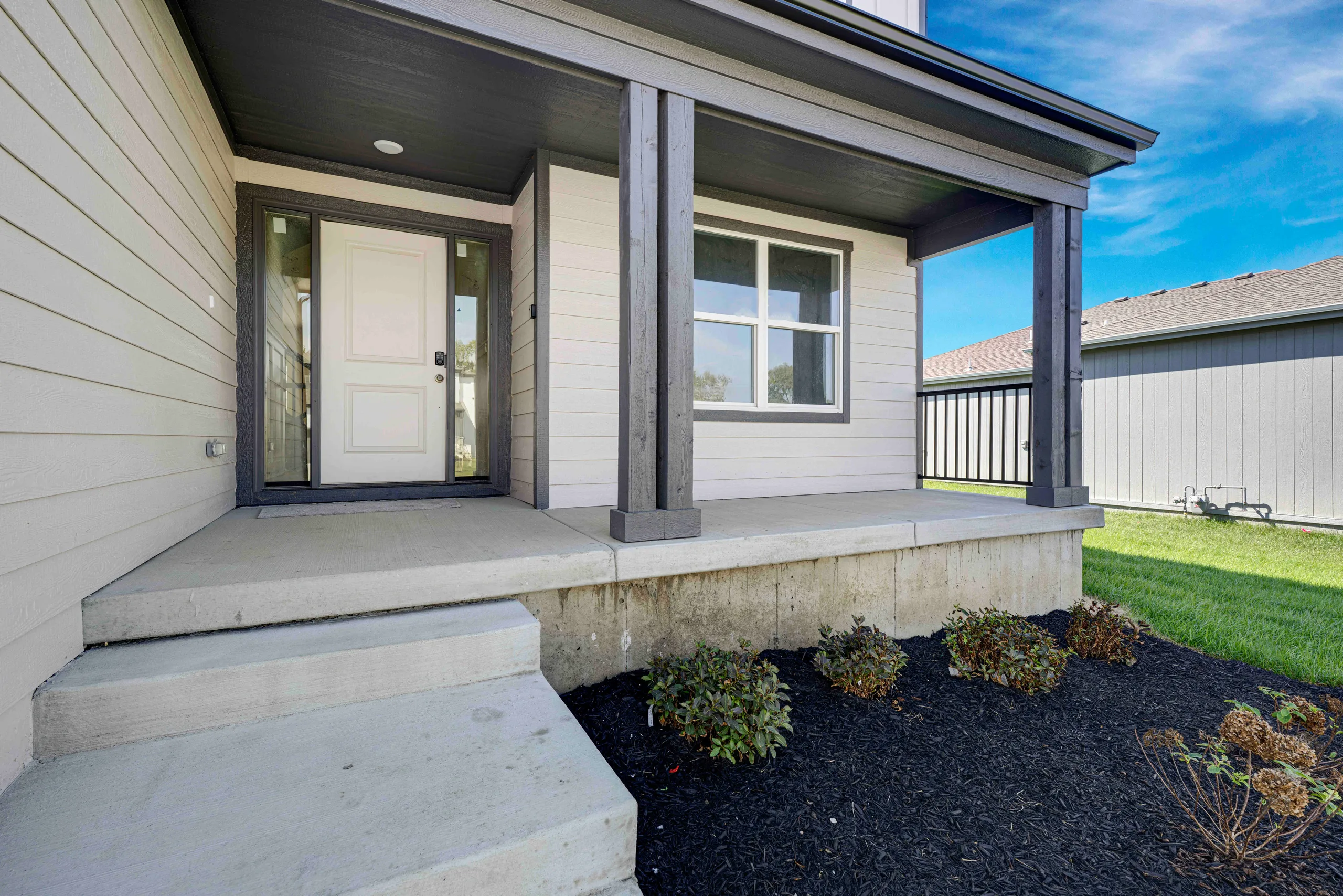 Front porch of a home with white door, dark beam overhang, concrete steps, and landscaped planting beds.