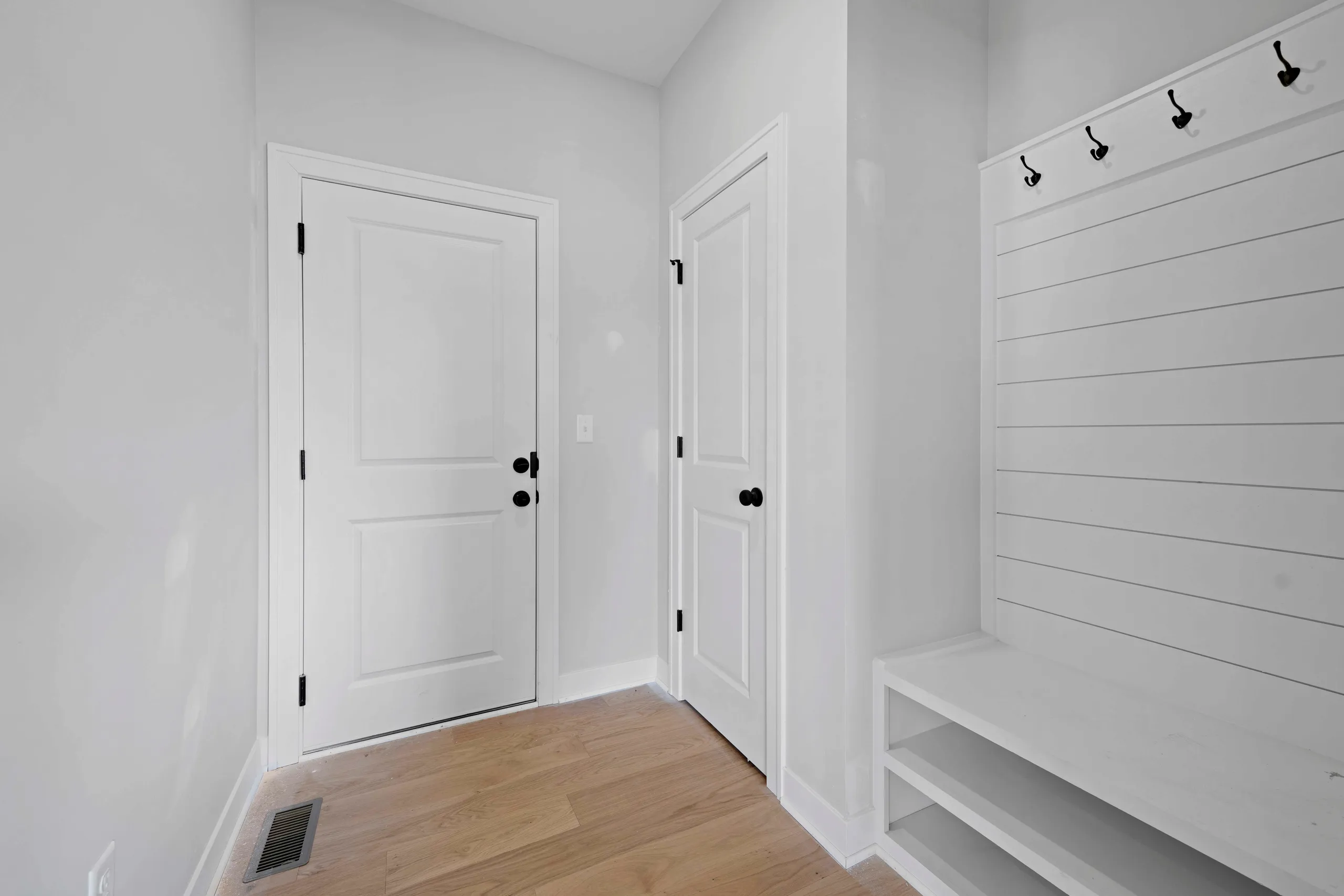 White mudroom with two white panel doors, shiplap wall with black hooks, and wooden flooring.