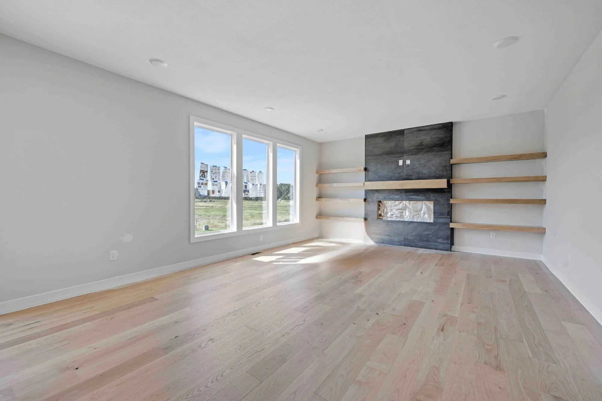 Empty living room with large windows, wood flooring, dark tile fireplace with wood shelving, and recessed lighting.