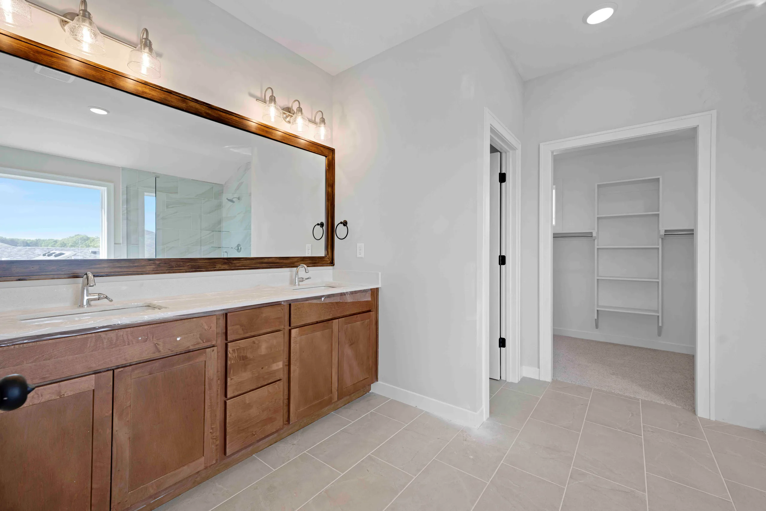 Bathroom with wood vanity, dual sinks, large mirror with lighting, and walk-in closet entrance.